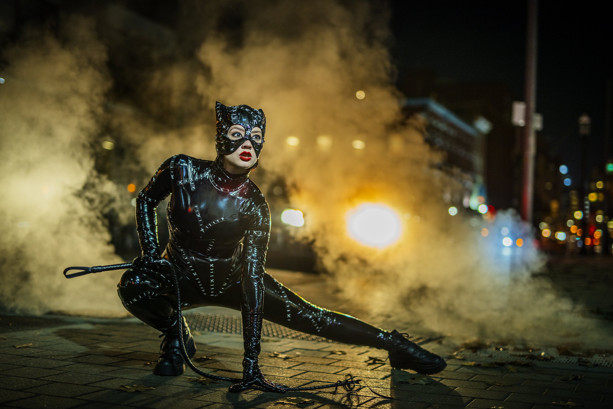 Woman dressed as Catwoman in a shiny black costume with a mask, posing with a whip on a city street at night with smoke and colorful lights in the background.
