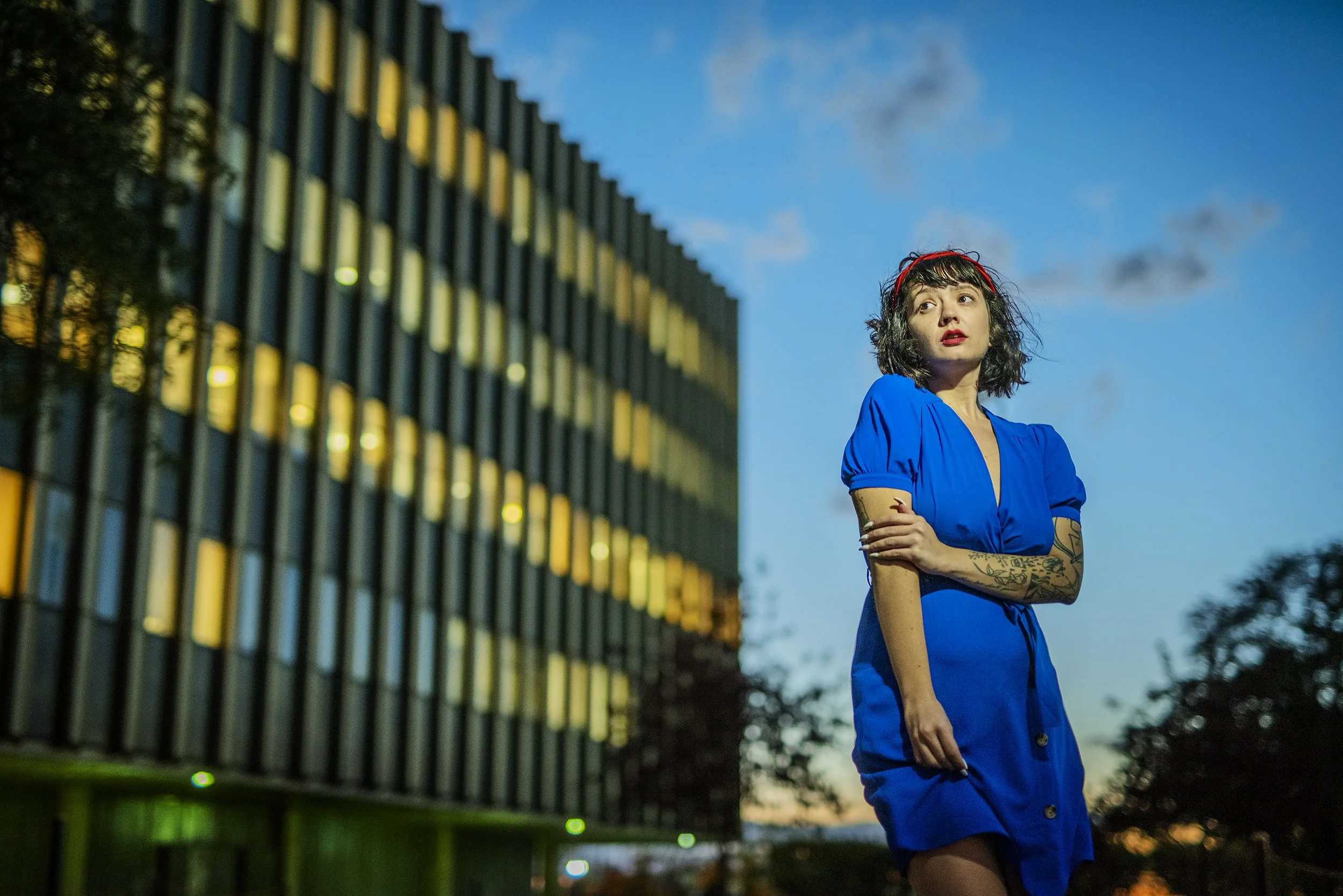 A woman with black curly hair, wearing a blue dress and red headband, standing outdoors with her arms crossed, during early evening with a modern building and cloudy sky in the background.