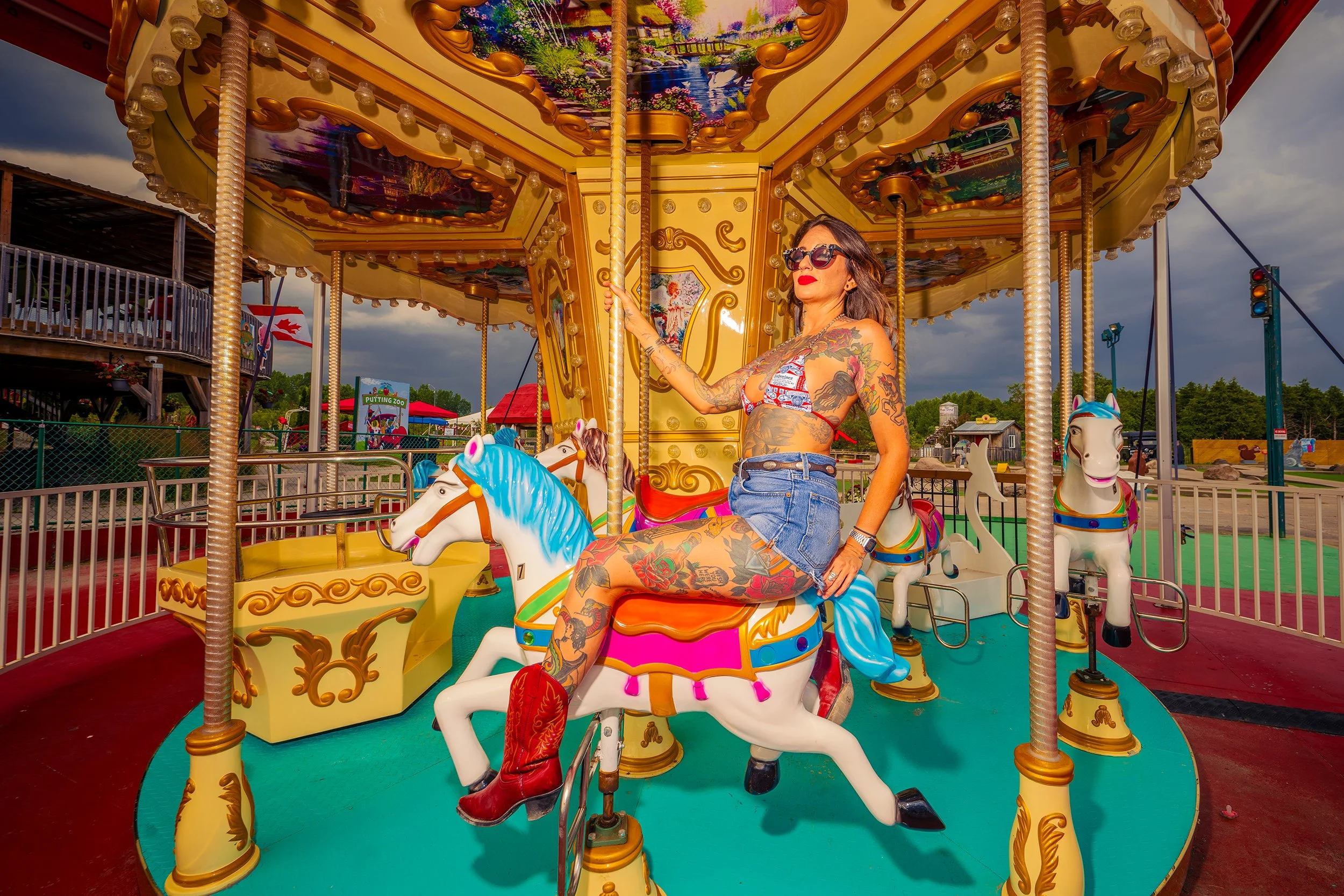 A tattooed woman wearing sunglasses, a red and white striped top, denim shorts, and red boots is sitting on a carousel horse at an amusement park during daytime with dark clouds in the sky.