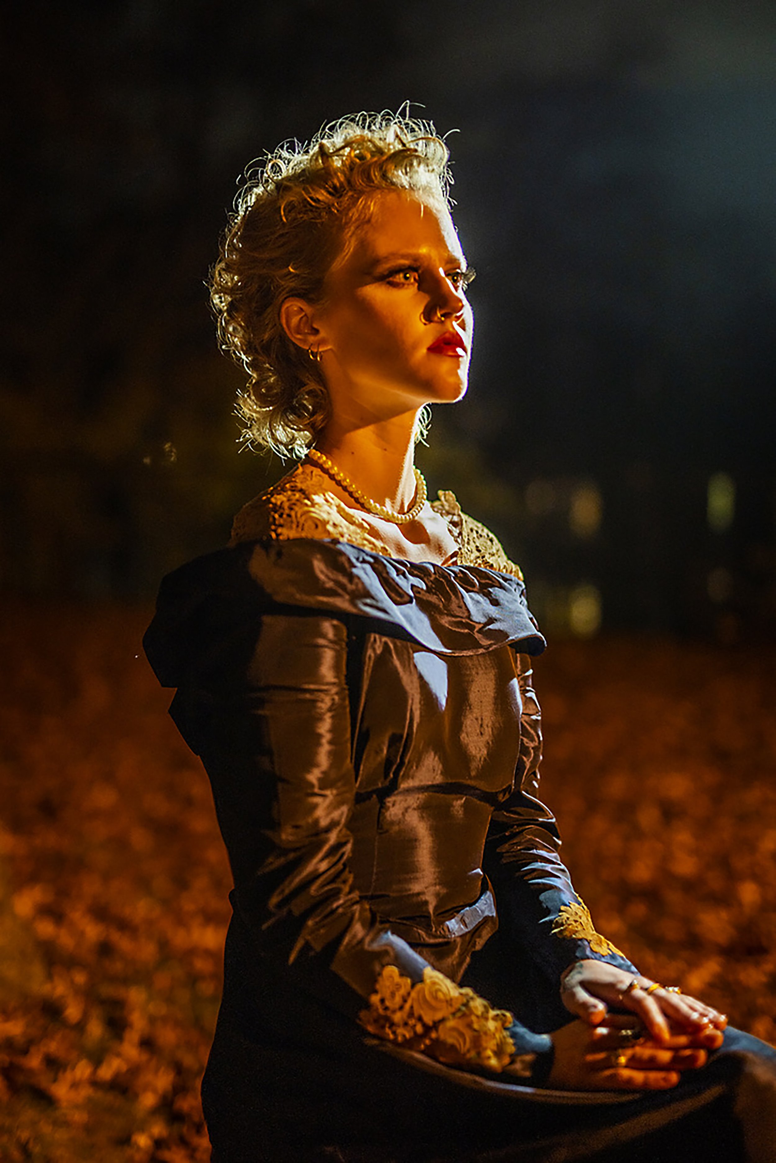A woman with curly blonde hair, wearing a vintage-style black dress with floral embroidery, seated outdoors at night with dim orange lighting and autumn leaves on the ground.