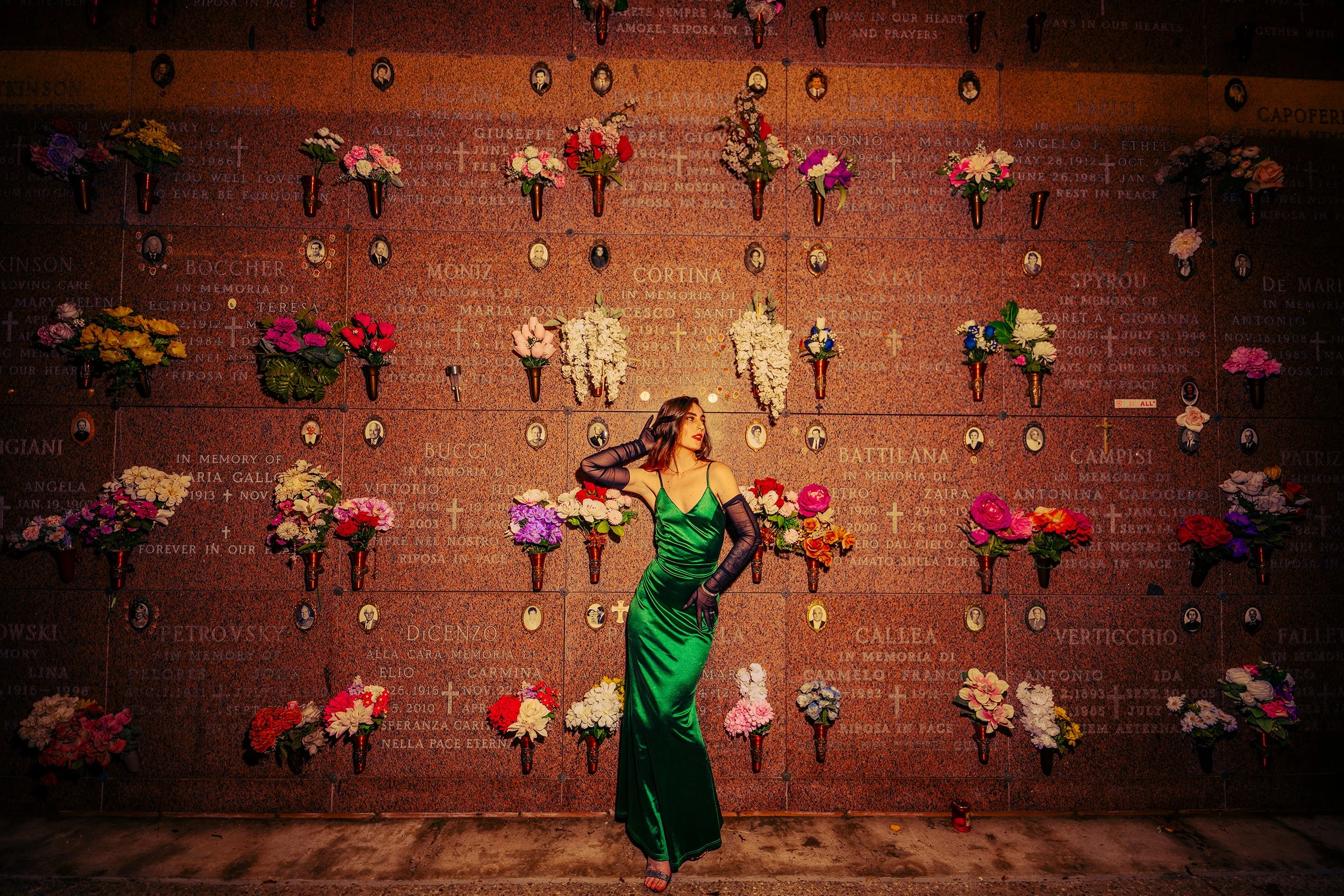 A woman in a green satin dress with black lace sleeves posing in front of a memorial wall decorated with colorful flowers and small portraits.