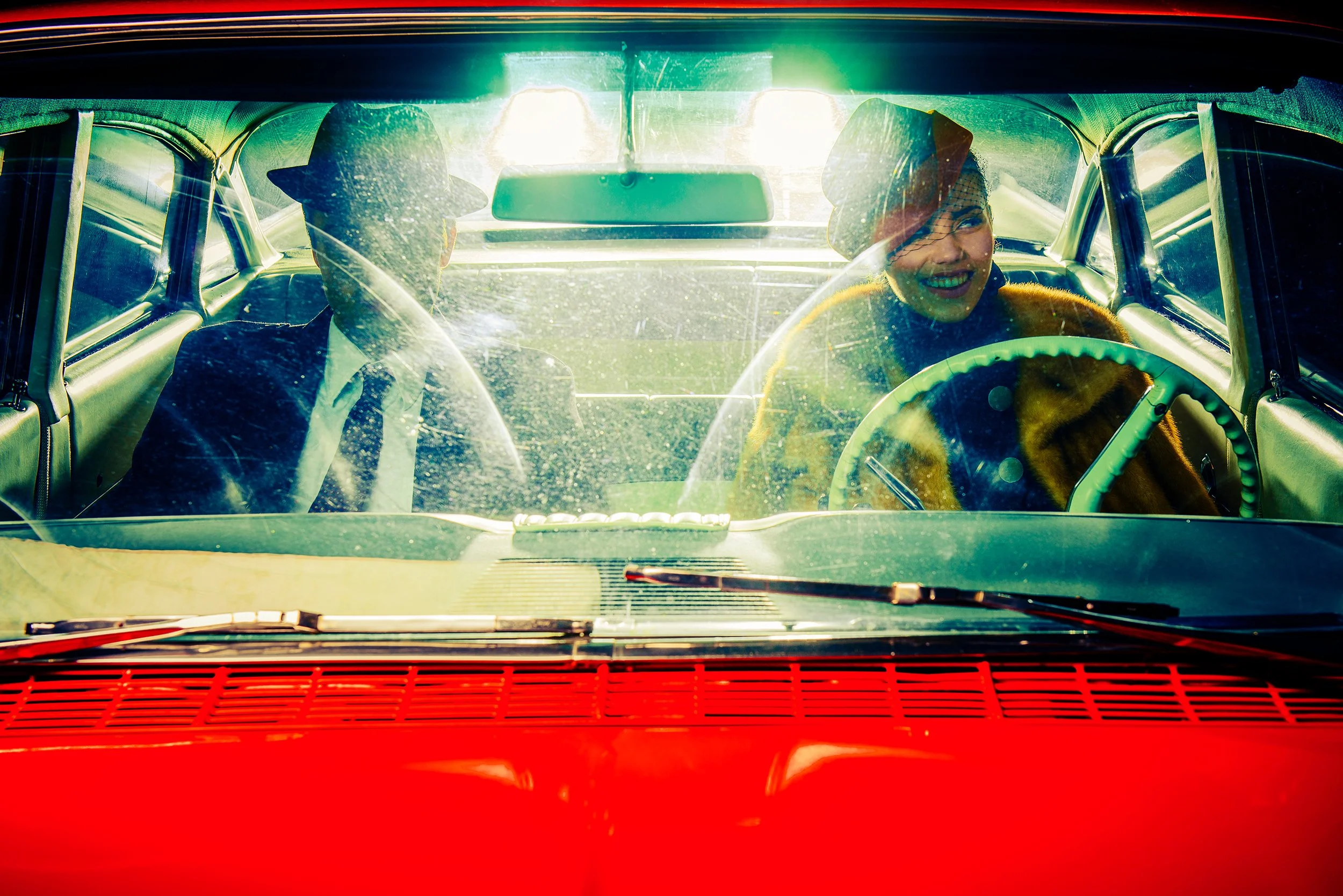 Inside a vintage car with two women wearing hats, one smiling, as seen through the windshield with sunlight shining in.
