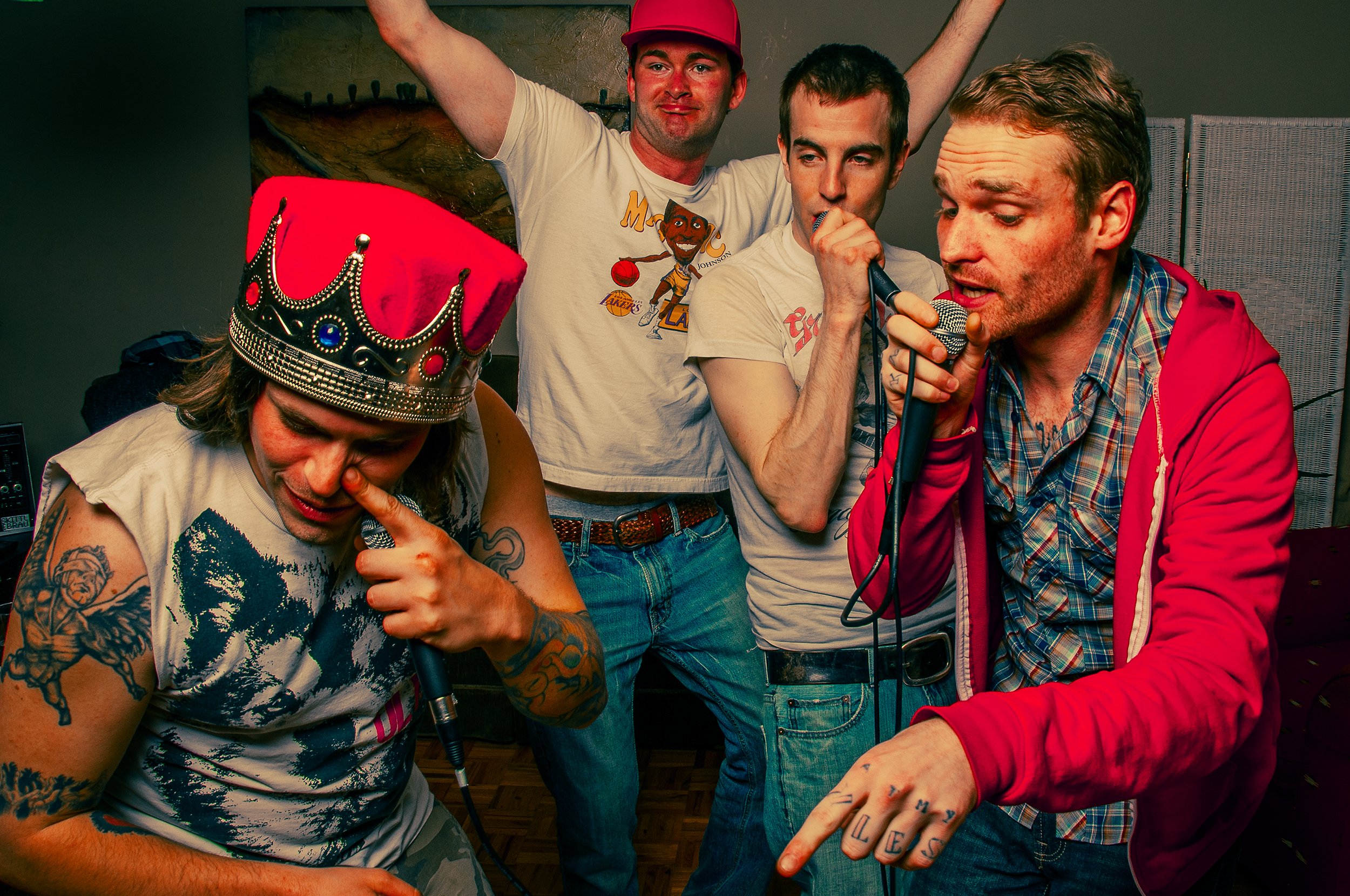 Four men are gathered closely with one woman who is wearing a red crown. They appear to be singing into microphones, with one man holding a microphone and another touching his lips.