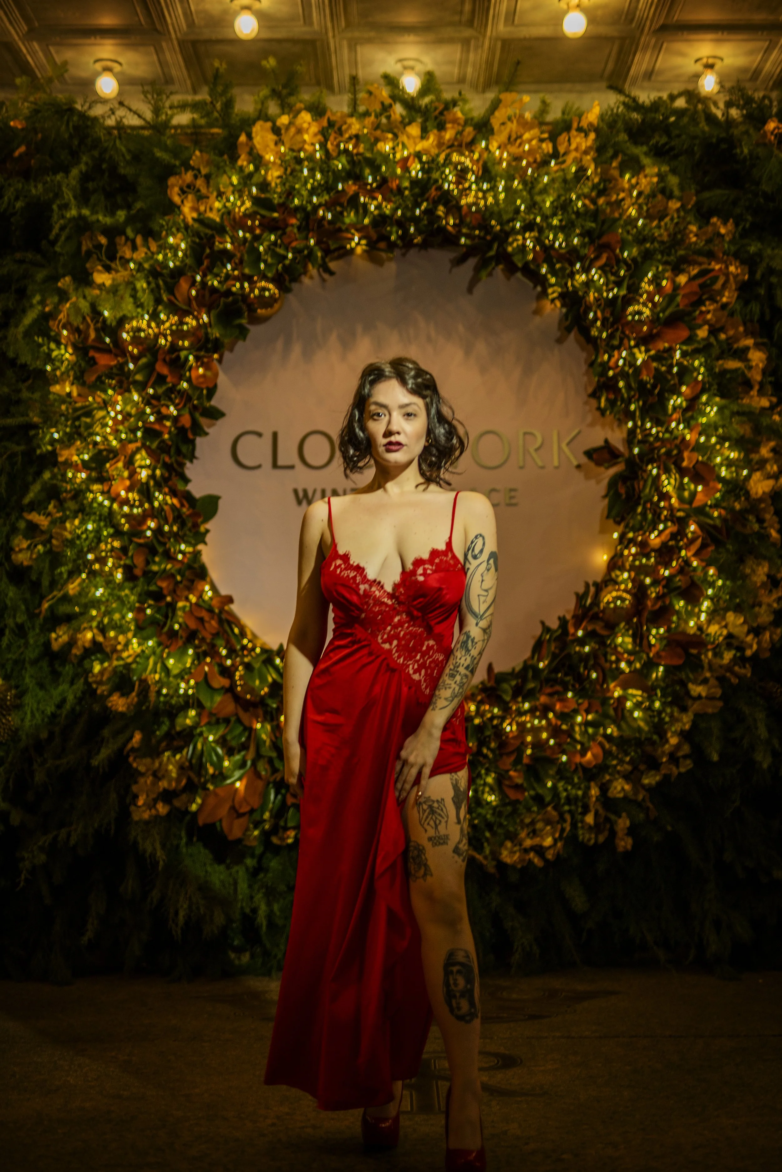 Woman in a red lace dress standing in front of a large floral wreath with lights, with the words 'CLOAK WORK' visible in the background.
