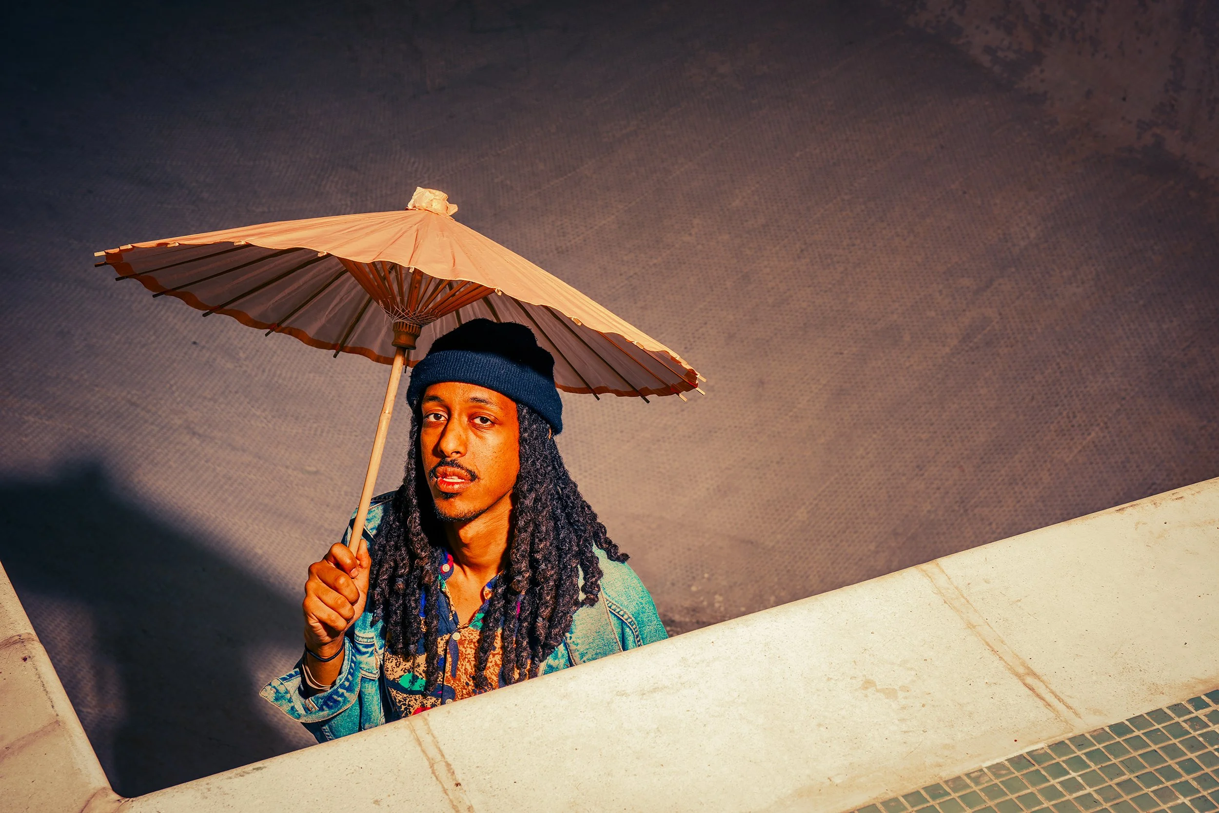 A man with long dreadlocks, wearing a beanie and colorful shirt, holding a pink parasol, standing near a hot tub or pool with a textured ceiling above.