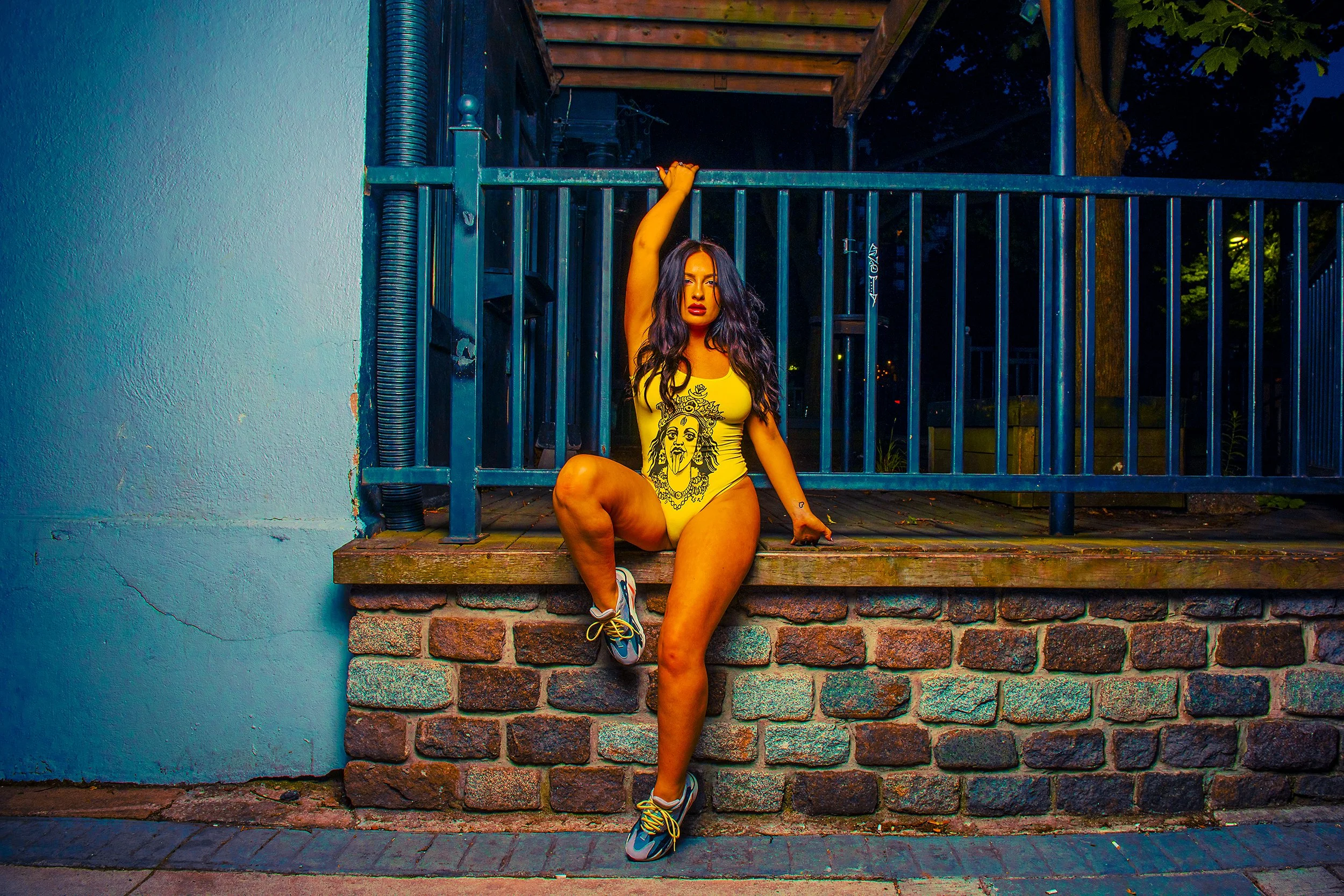 A woman in a yellow tank top with a graphic print, yellow shorts, and running shoes is posing on a brick ledge in an urban setting at dusk. She is sitting with one leg bent and the other extended, with one arm raised and holding onto a railing above 