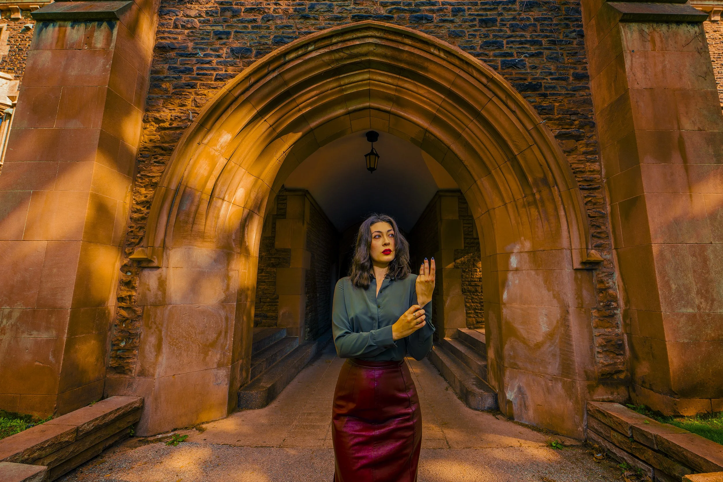 A young woman with dark wavy hair and red lipstick standing under a brick archway, looking to her left, with her right hand raised and pointing, in front of an old stone building with a hanging lantern.