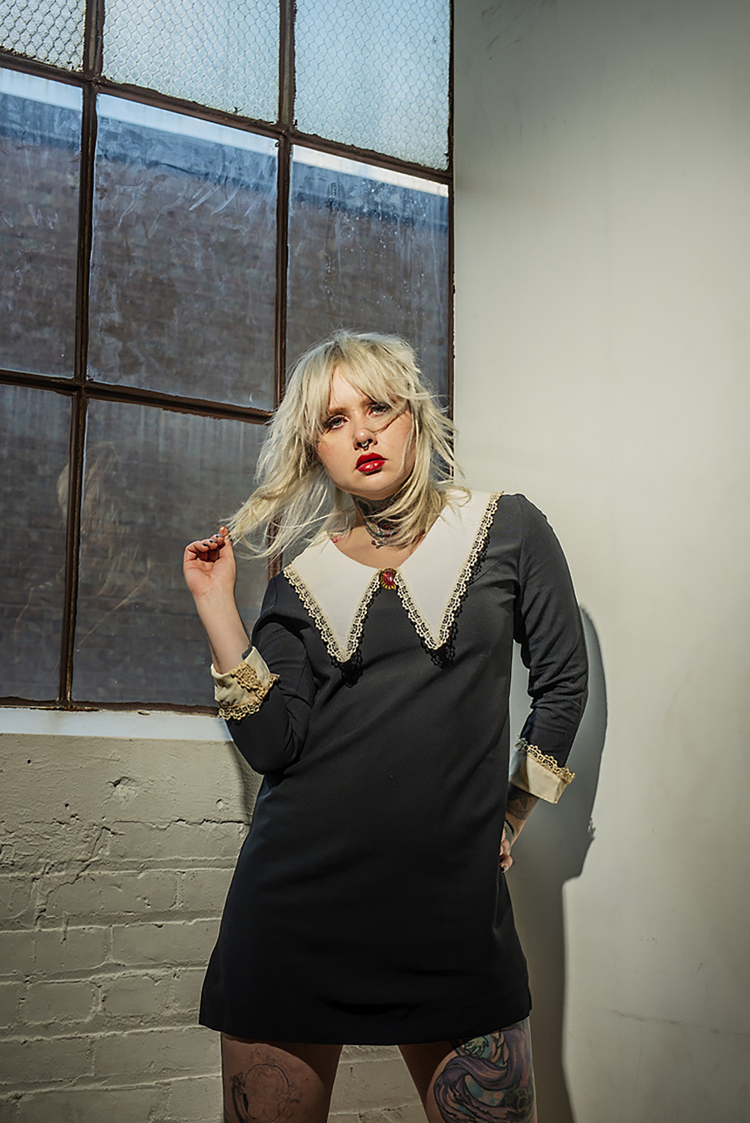 A woman with platinum blonde hair, wearing a black dress with beige lace trim, standing indoors near a window with rusty brown framing. She has tattoos on her legs, and is looking at the camera with her hand touching her hair.
