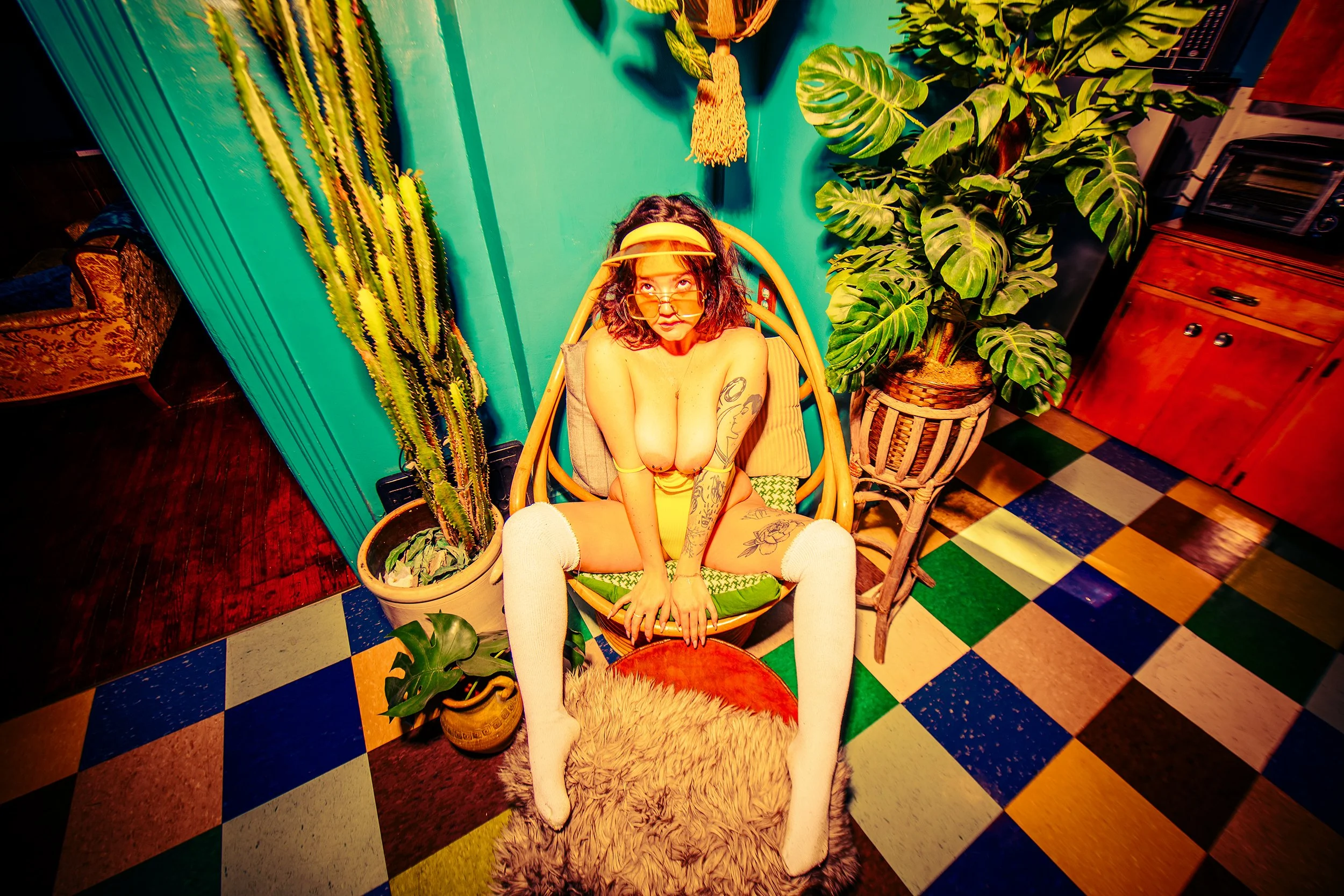 A woman with glasses and tattoos sitting on a chair with crossed legs, wearing knee-high socks, in a room with vivid green walls and colorful tile floor. Surrounding her are various green plants and a cactus, with a wooden cabinet and toaster on the 