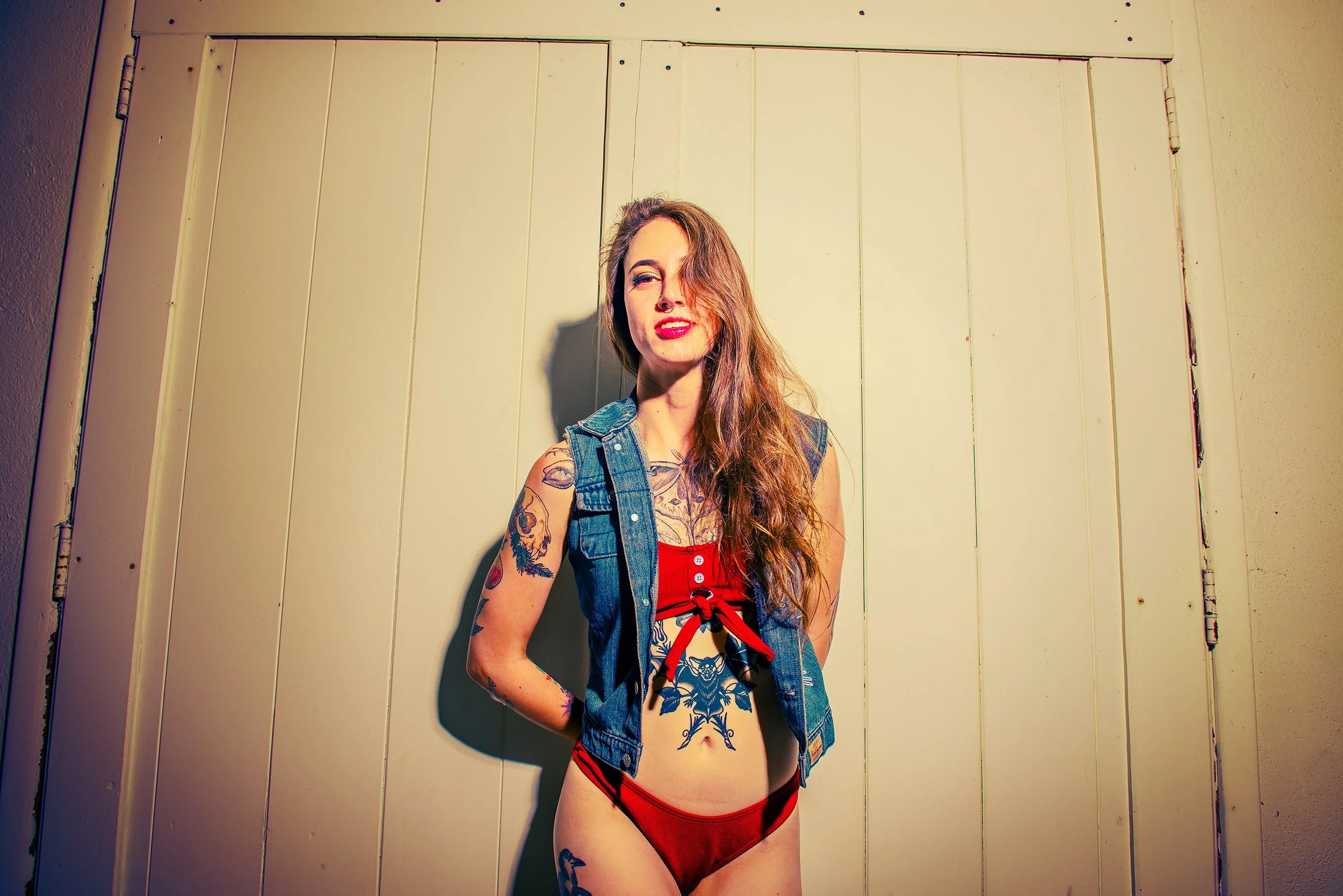 A woman with long brown hair and multiple tattoos stands against a beige wooden wall, wearing a sleeveless denim vest over a red bikini top and matching bottom, with her left hand on her hip, smiling and squinting in the sunlight.