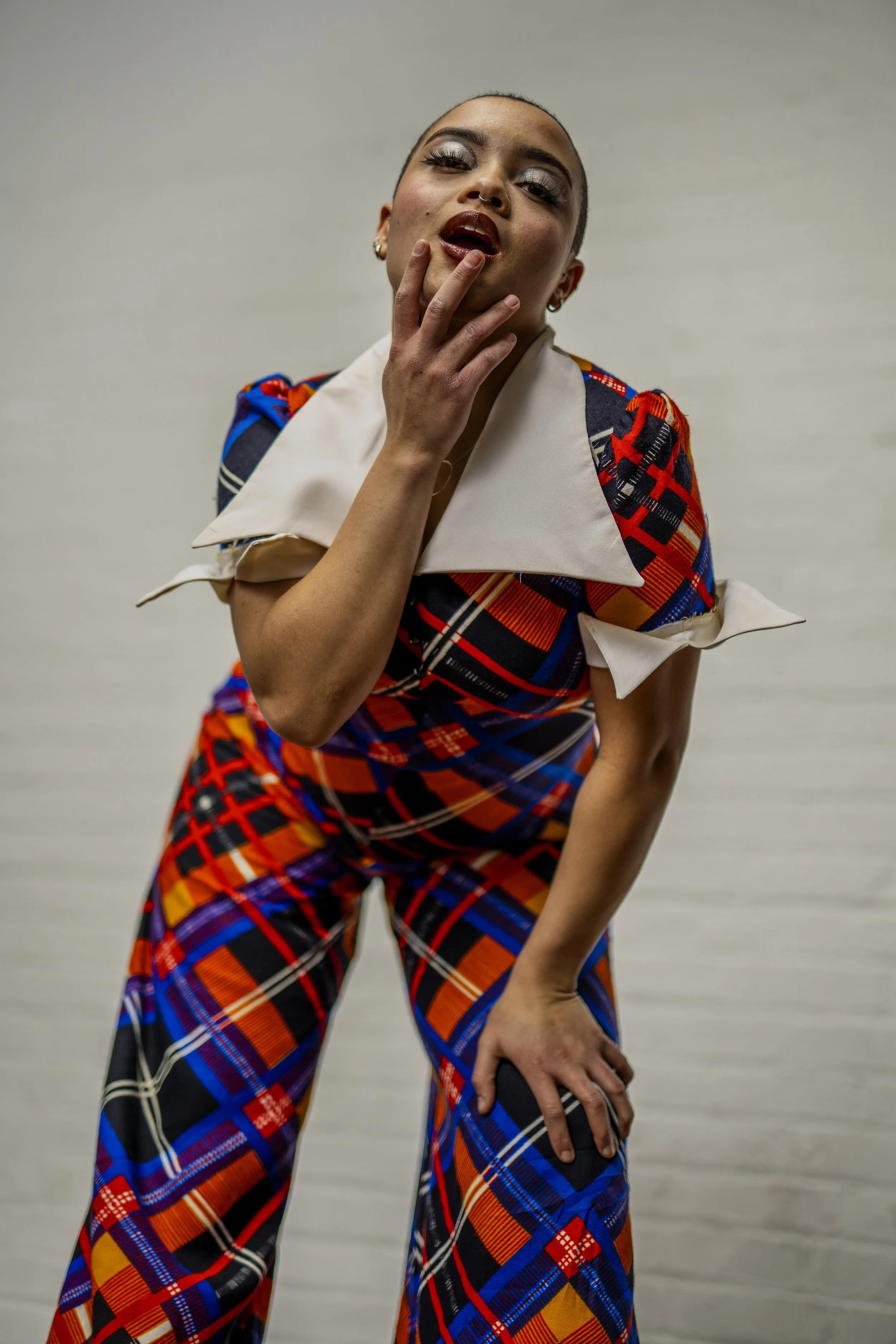 A person wearing bold, colorful plaid clothing, striking a confident pose with one hand on their thigh and the other touching their chin, with a plain neutral background.