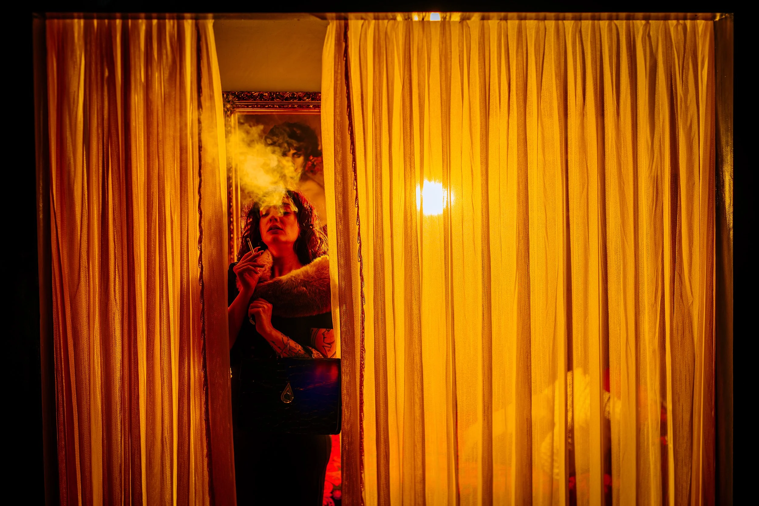 A woman stands behind yellow curtains, smoking. There is a mirror reflecting her face and a framed picture of a person in the background, with warm orange lighting.