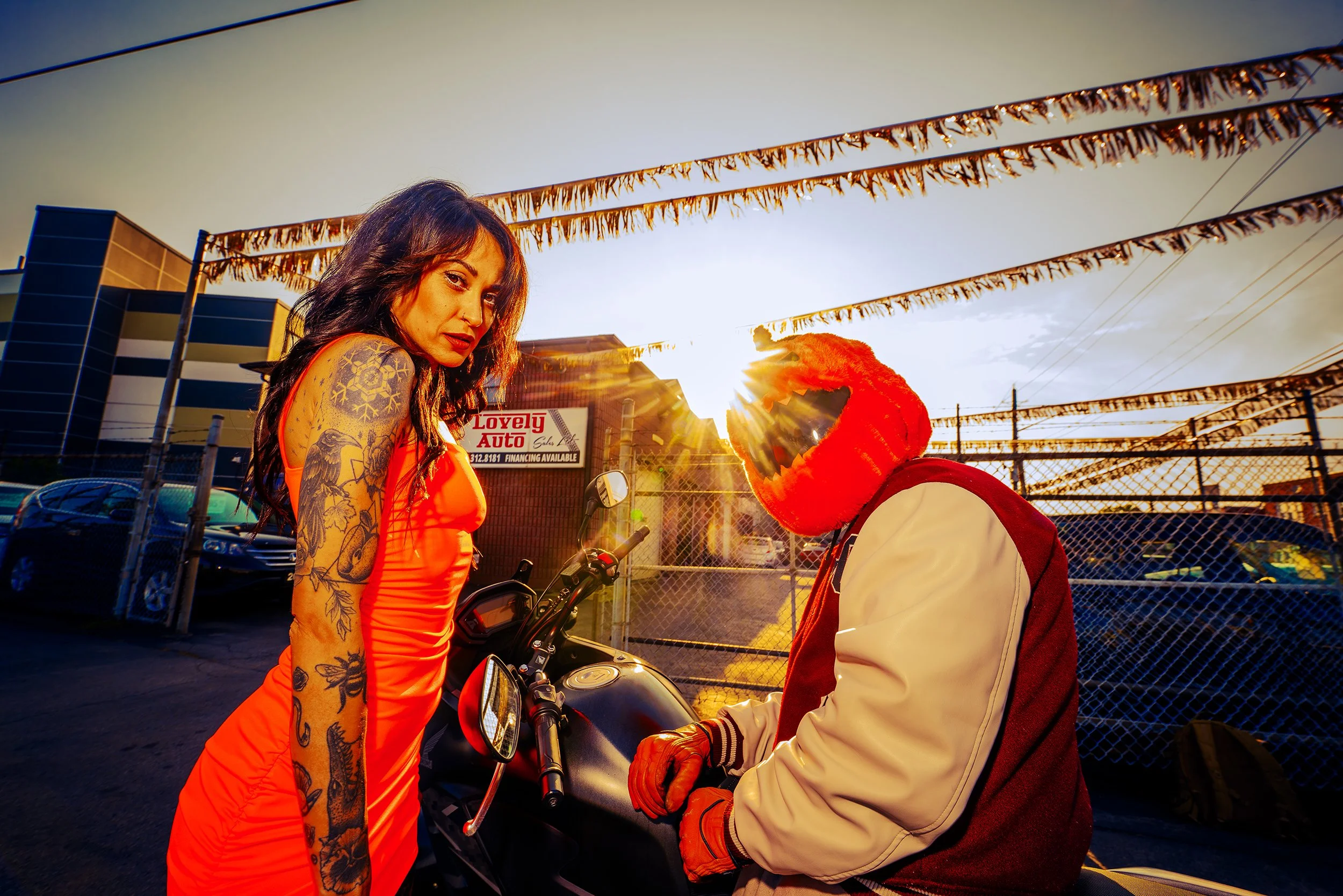 A woman with tattoos on her arm and dark hair, wearing a red dress, stands beside a person in a red and white jacket and red gloves, who is sitting on a motorcycle during sunset. The background features a chain-link fence, flags, and a building with 