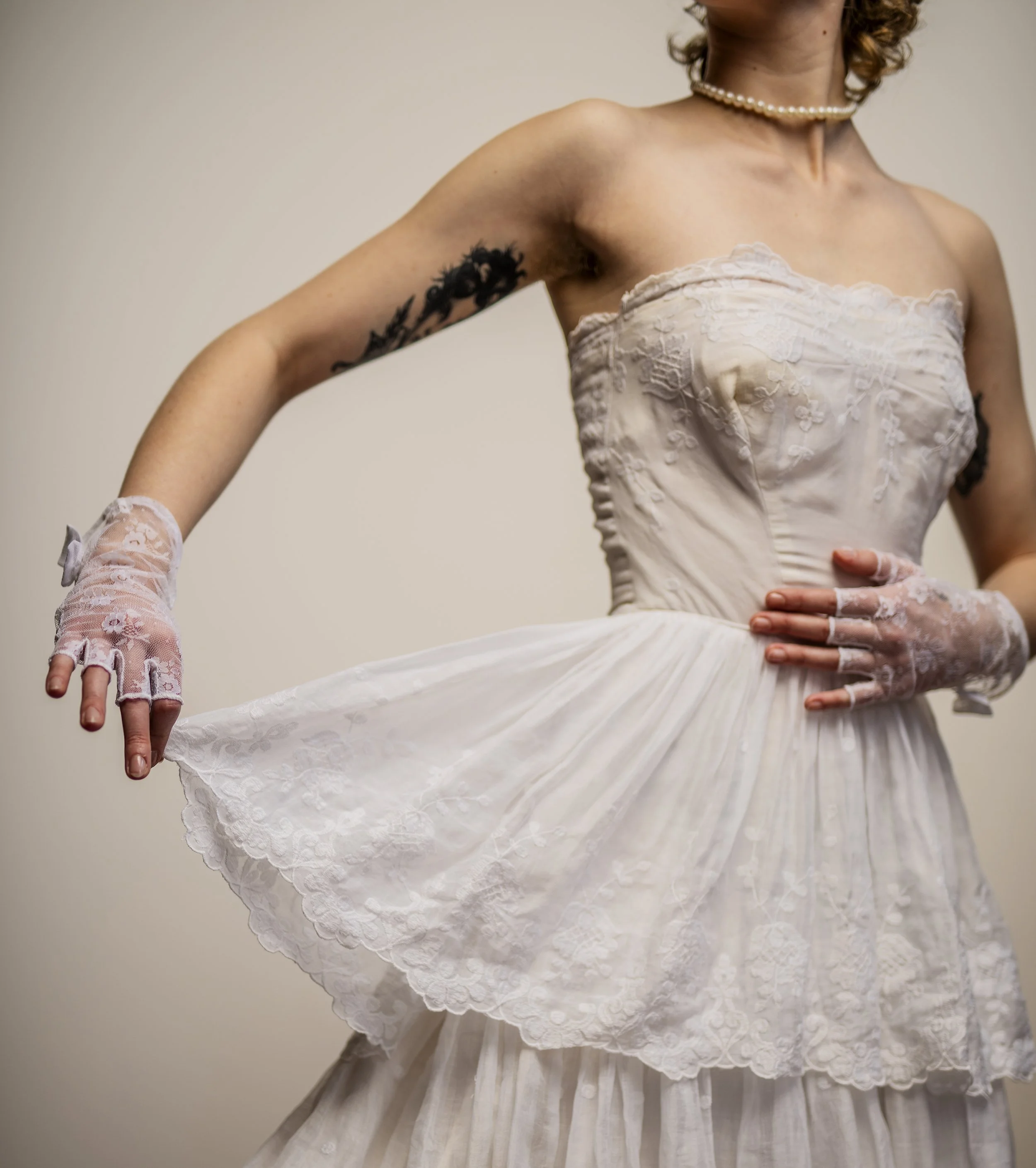 A woman is wearing a strapless white lace wedding gown with a floral pattern, pearl necklace, lace fingerless gloves, and has a tattoo of a floral design on her upper left arm, posing against a plain neutral background.