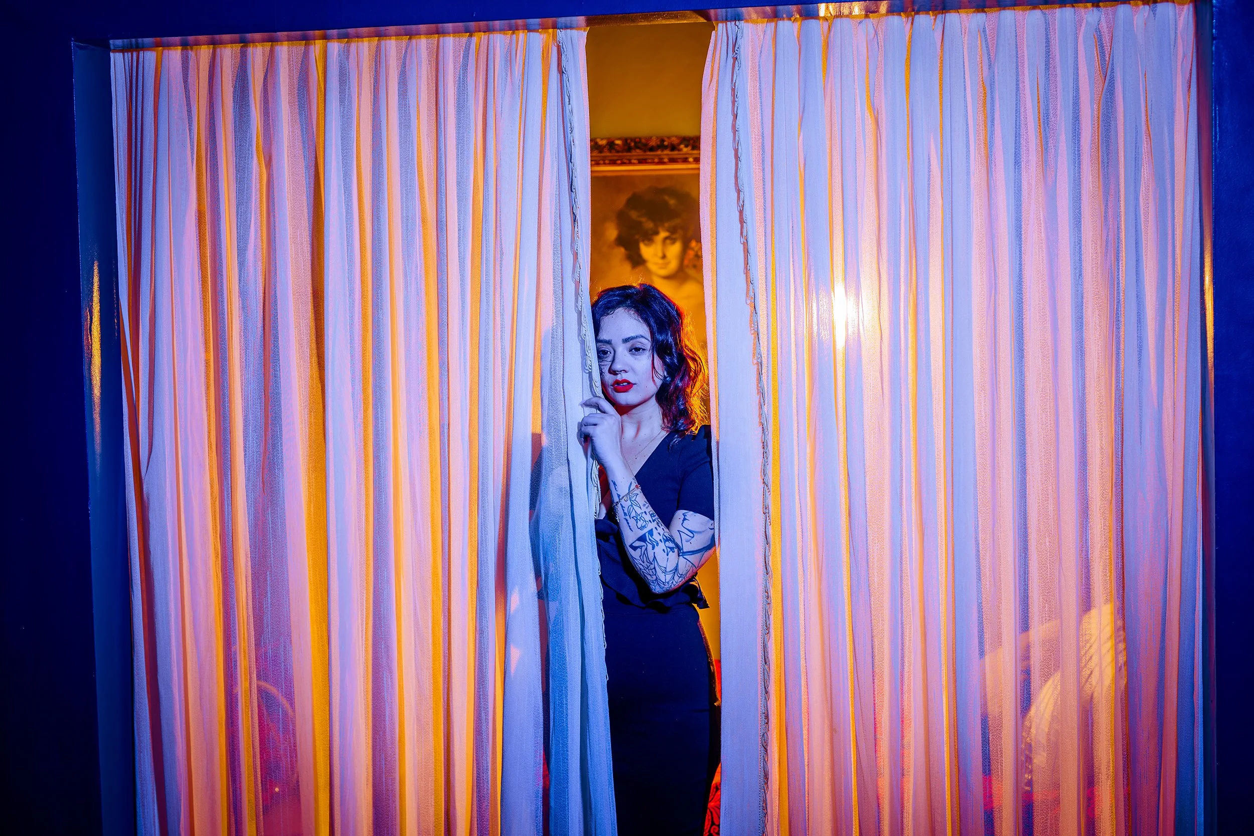 A woman with dark hair and tattoos on her arm peeks through a gap in multicolored curtains, with a portrait of a woman with curly hair visible in the background.