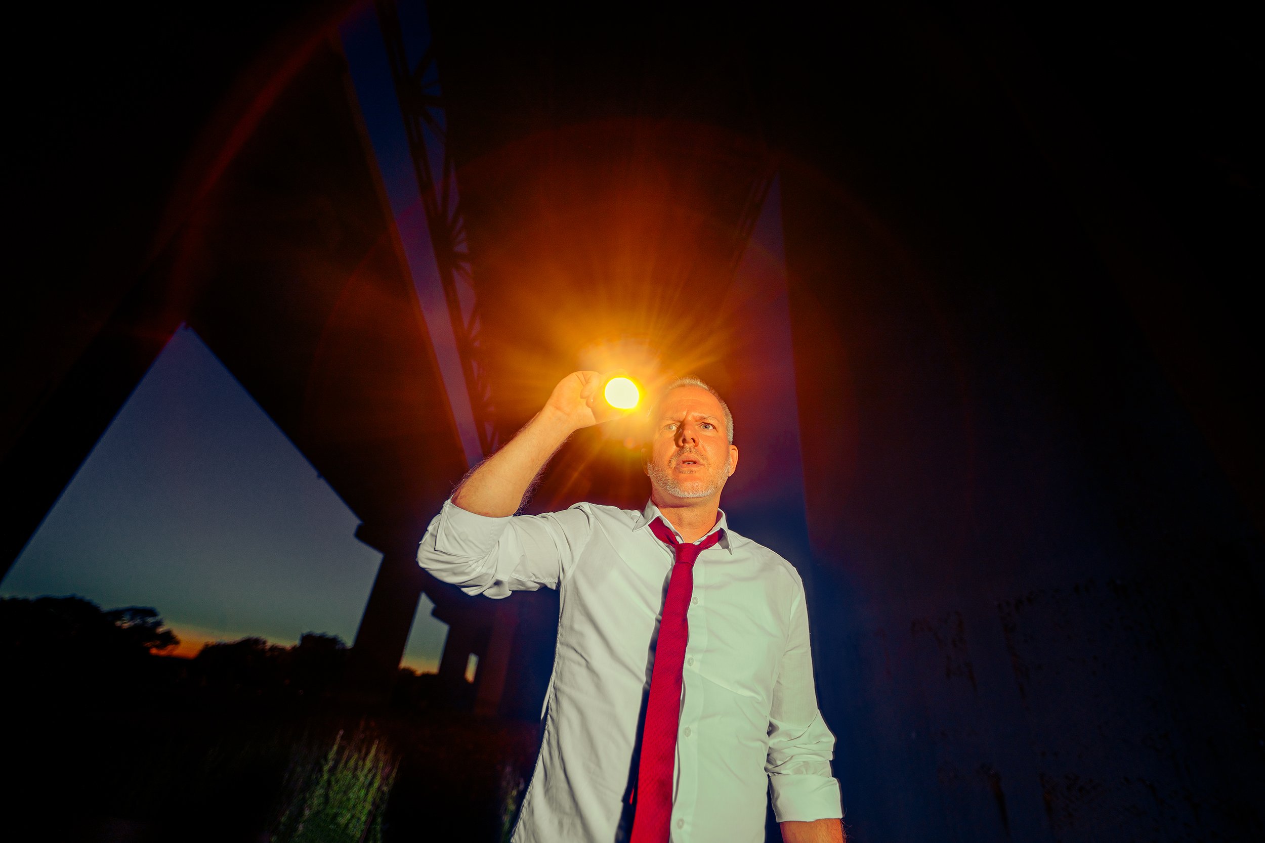 A man in a white shirt and red tie holds a bright yellow light at sunset, standing under a large bridge with the sky darkening behind him.