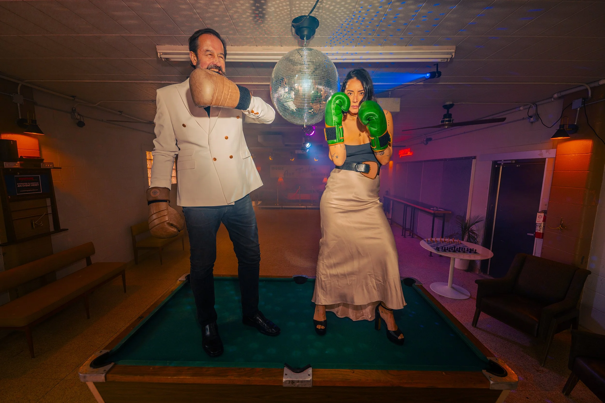 A man wearing boxing gloves and a woman wearing boxing gloves posing on a pool table with a disco ball overhead in a dimly lit room with colorful lights.