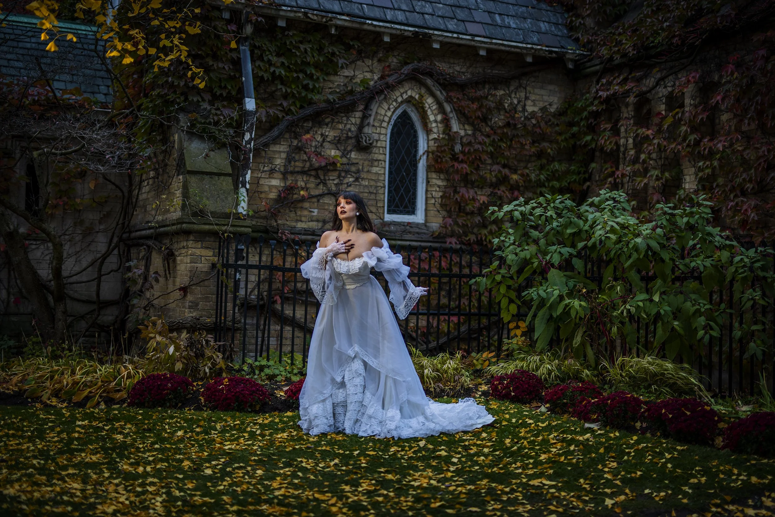 A woman in a vintage white wedding dress standing outdoors in a garden surrounded by fall foliage and flowers, with a brick building and ivy climbing on the walls in the background.