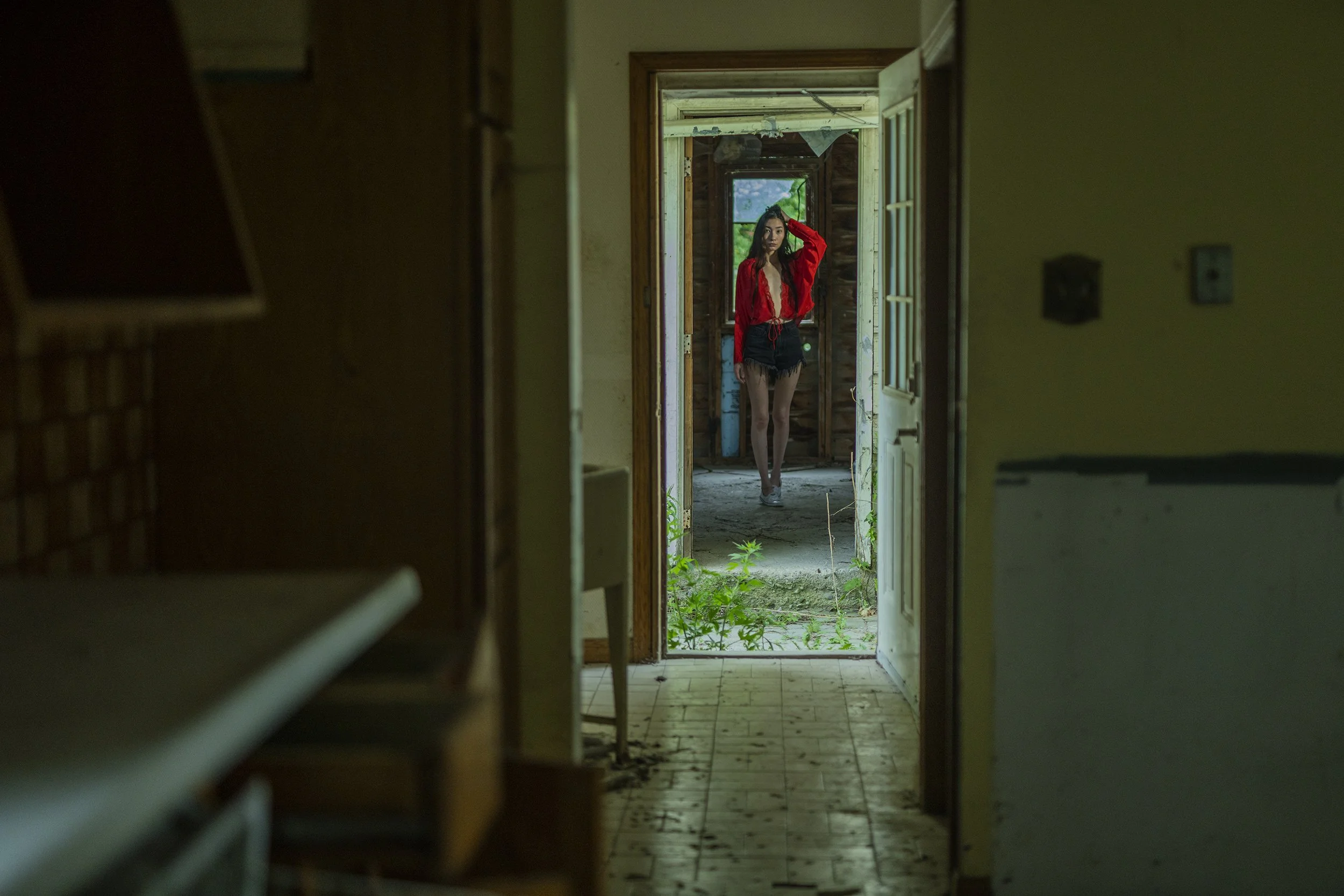 A young woman in a red top and black shorts standing in a hallway of an abandoned house, looking directly at the camera.