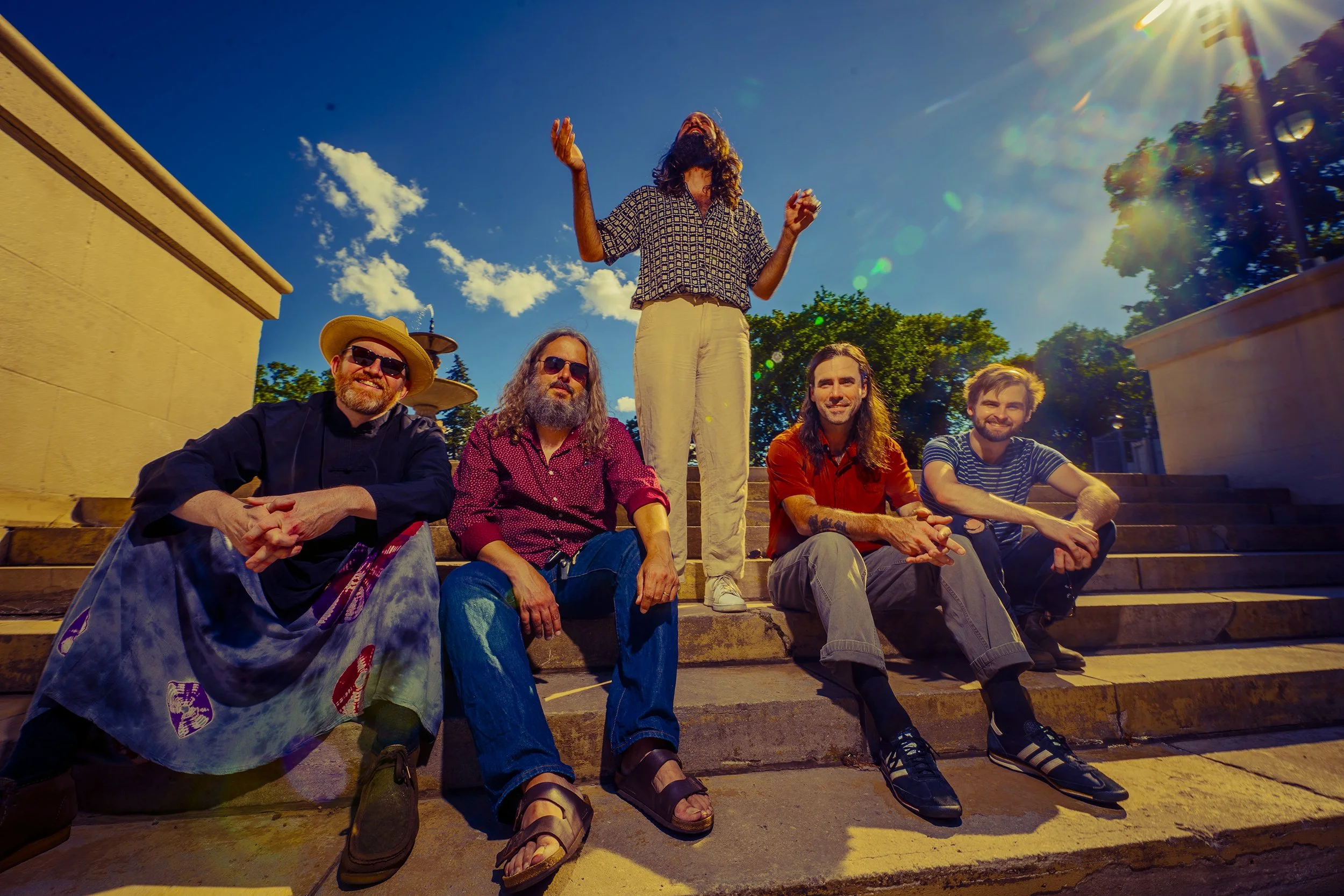 A group of five friends sitting on outdoor steps under a bright blue sky, with one standing behind them. The group is diverse, dressed casually, enjoying the sunny weather and smiling.