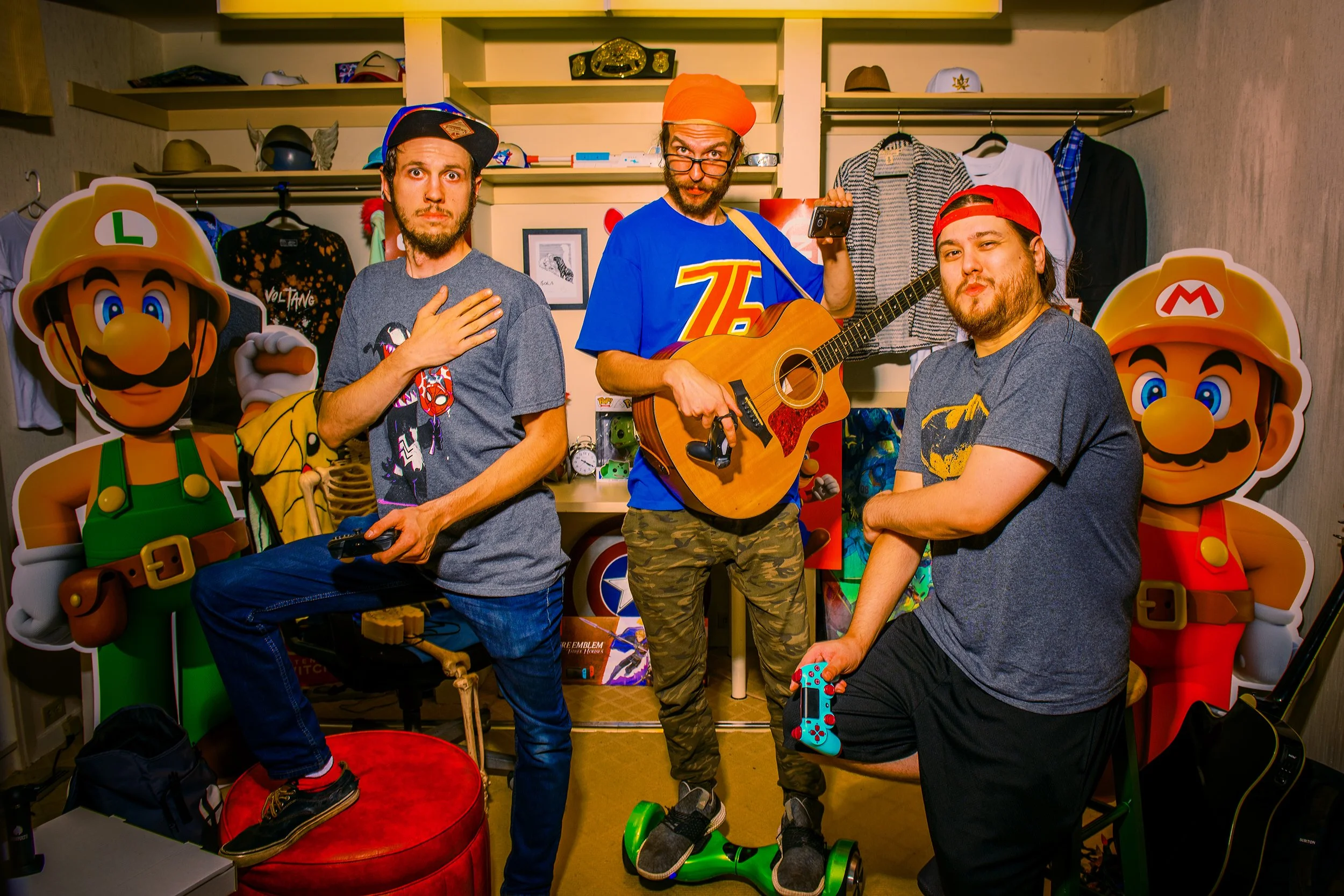 Three men in a room filled with video game and pop culture memorabilia. Two are standing, one sitting, each holding a gaming controller. The middle man is wearing a blue shirt with a large yellow and red logo and a red beanie, playing a guitar. The m