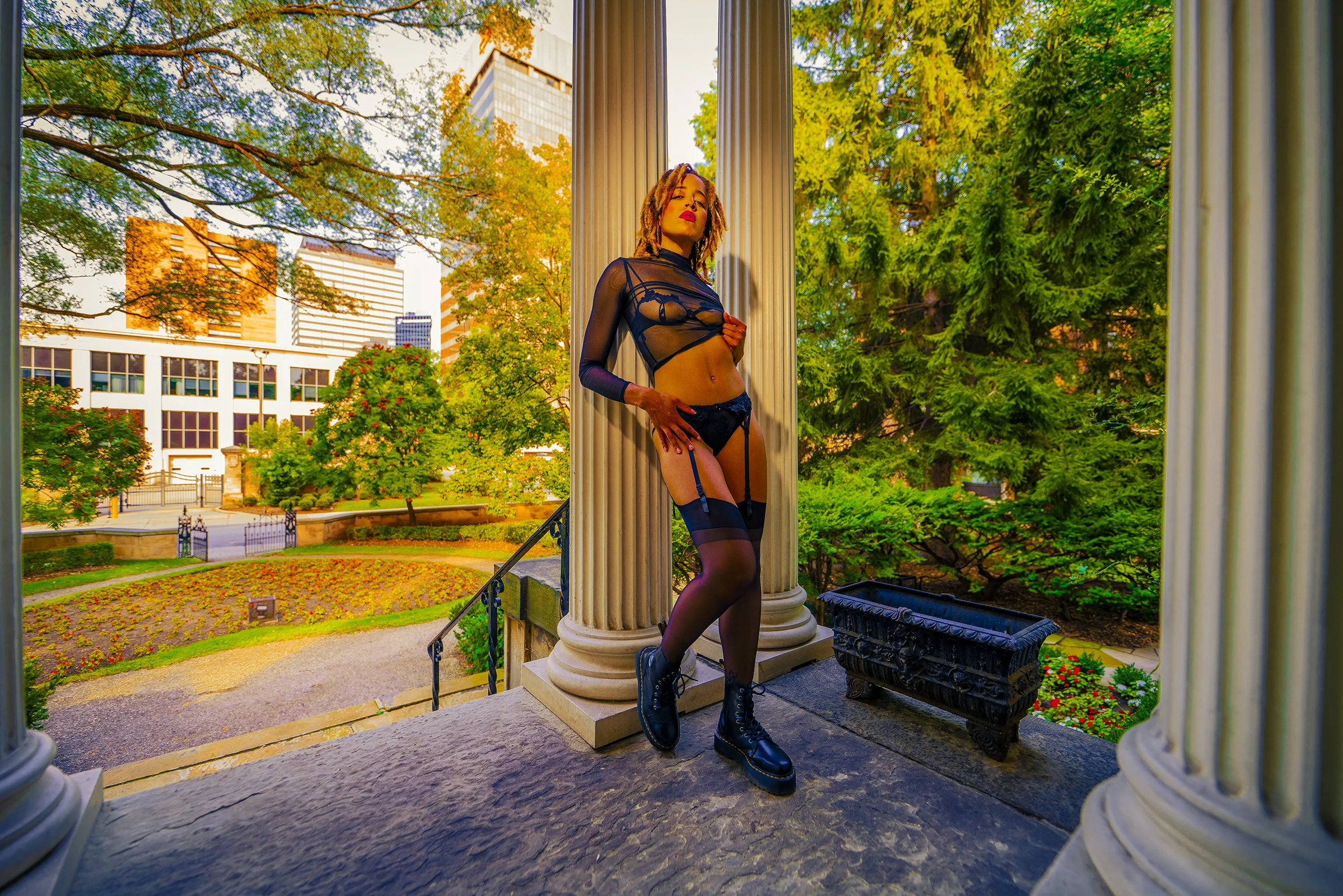 A person stands between two large white columns on the steps of a building, wearing black lingerie with sheer, patterned stockings and combat boots. The person holds their chest with one hand and places the other on their hip, with an urban landscape