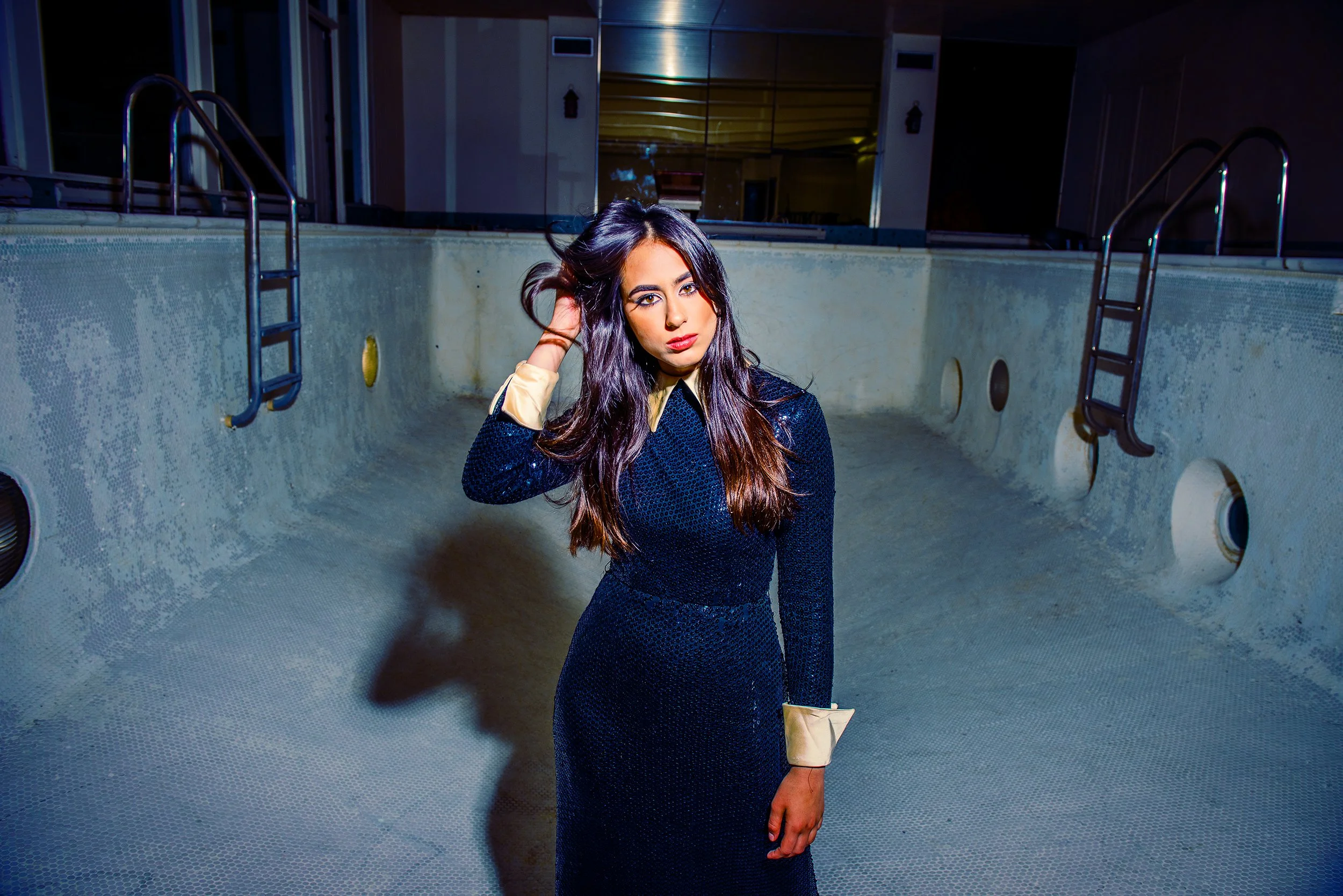 A woman with long dark hair and wearing a dark blue sparkly dress stands in an empty indoor pool area at night, touching her hair with her right hand and looking at the camera.