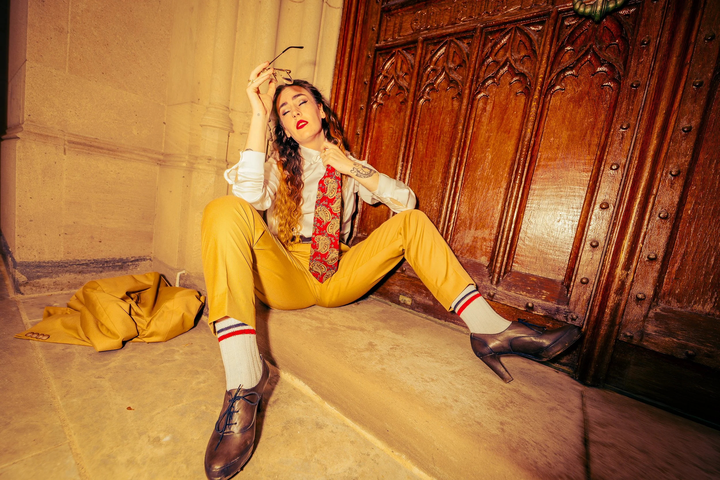 A woman with long curly hair, dressed in a white blouse, red patterned tie, and yellow pants, sitting on the floor against a wooden door with her eyes closed and a relaxed expression. She is holding glasses in her right hand and has a tattoo on her l