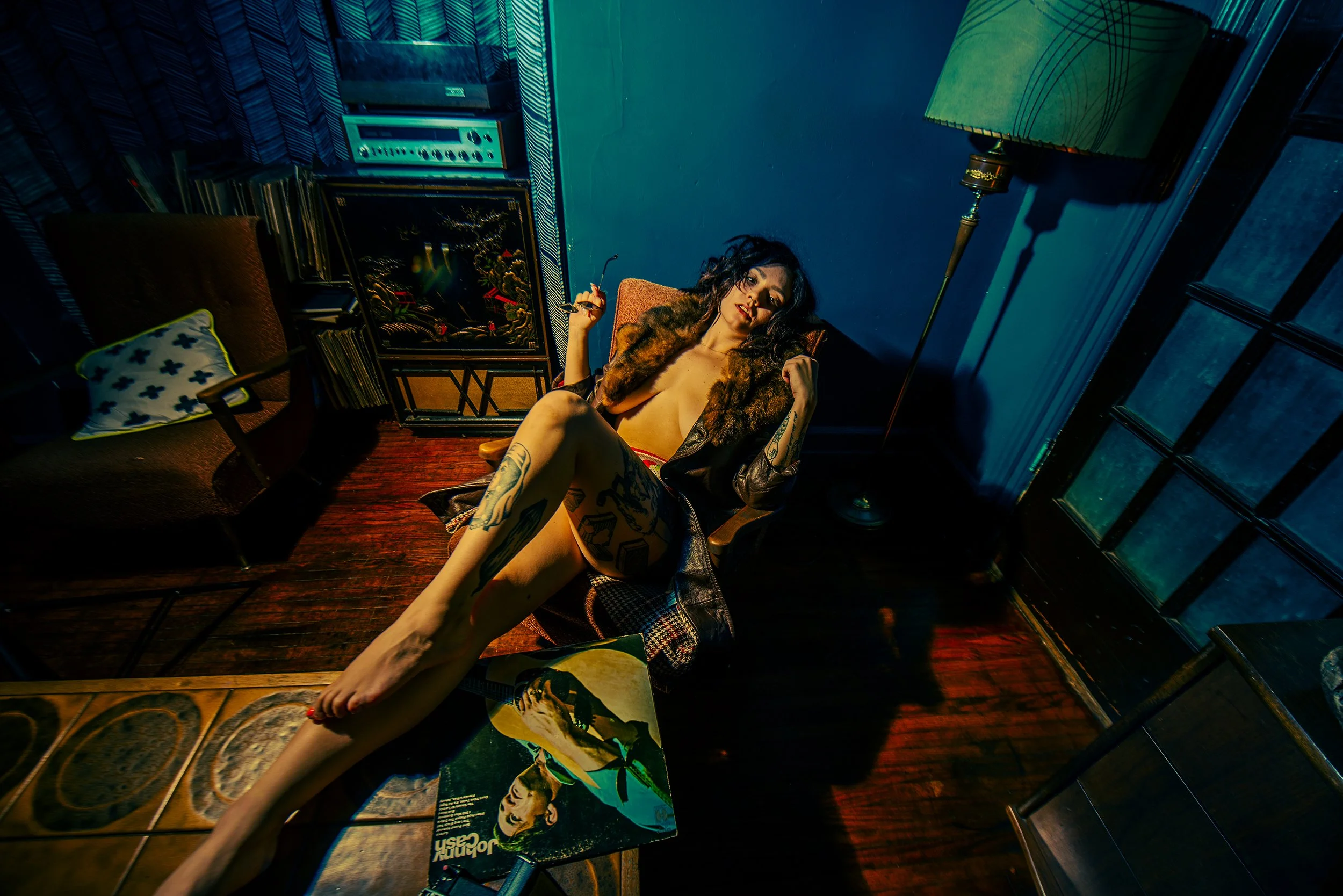 A woman with dark curly hair, tattoos on her legs, and wearing an open fur coat, lying on a chair in a dimly lit room with vintage furniture and artwork.