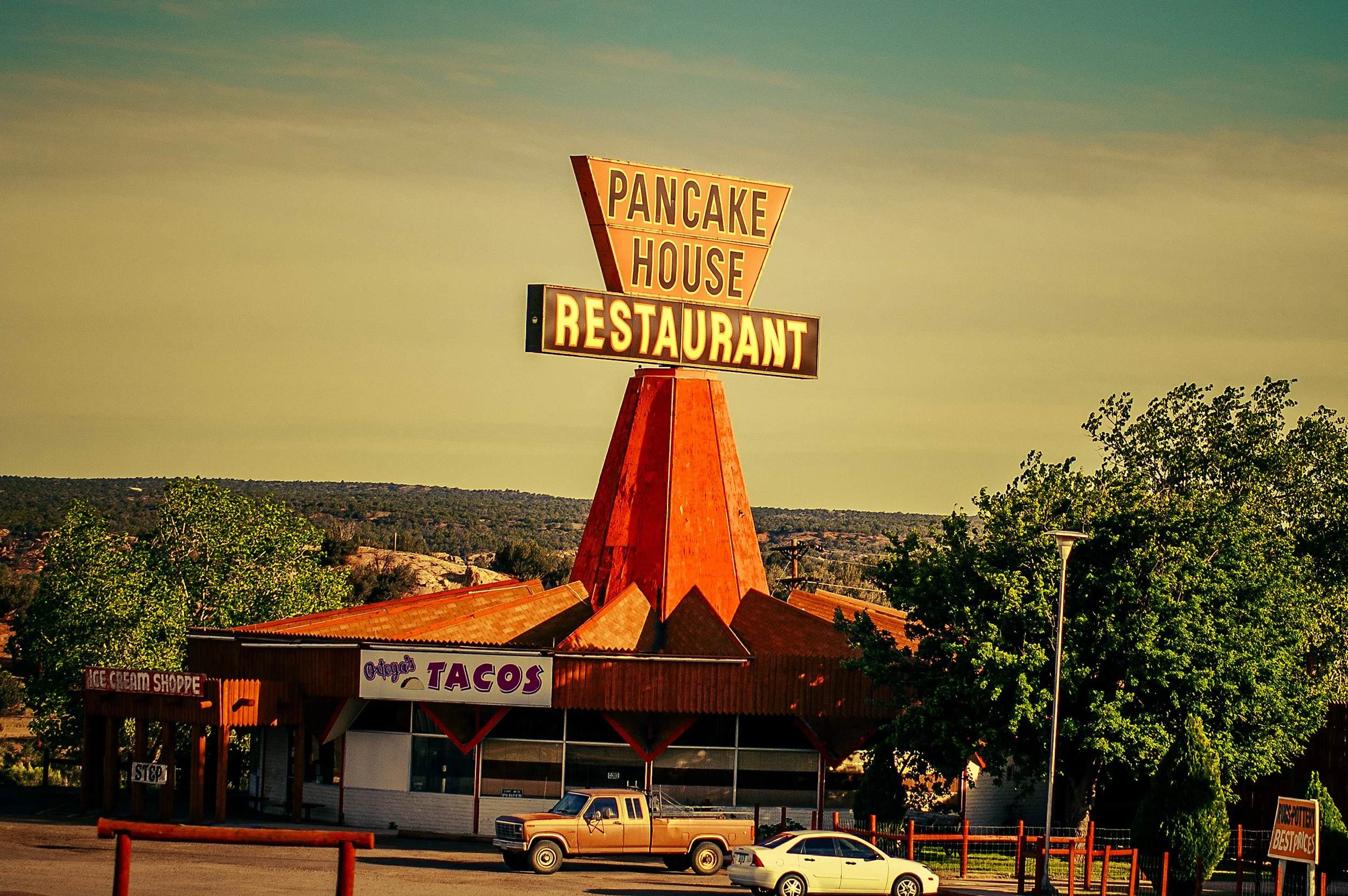 Sign reading 'Pancake House Restaurant' on top of a building, with a parking lot and cars in front, surrounded by trees and a clear sky.