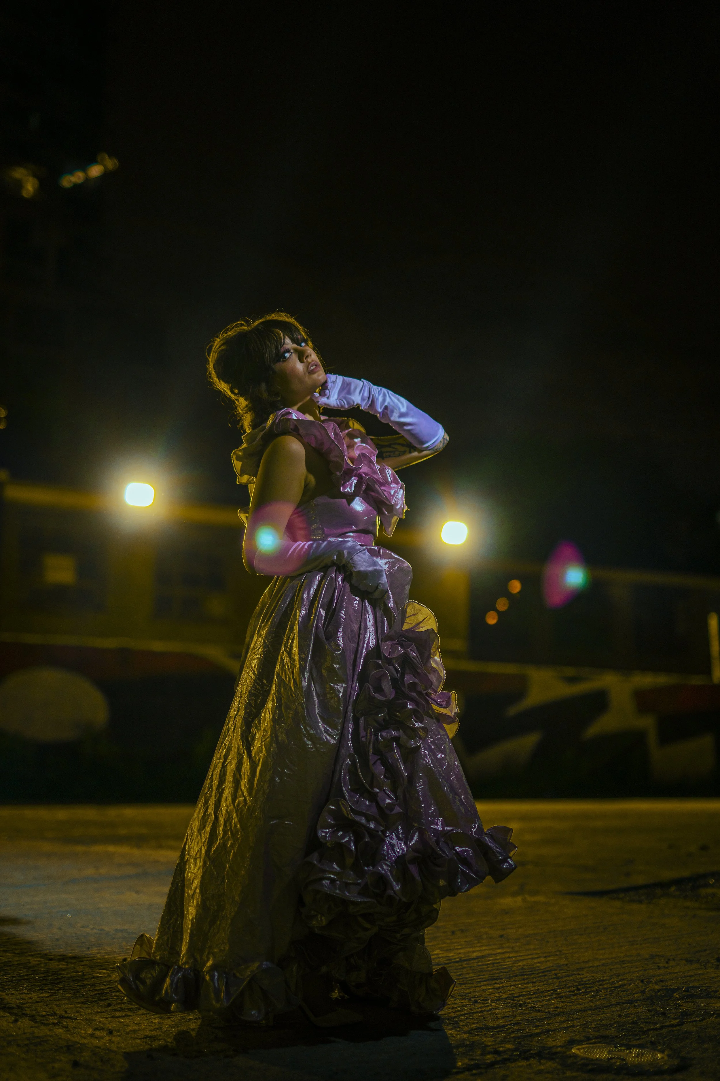 Woman in elaborate purple gown and long white glove standing on city street at night, illuminated by streetlights.