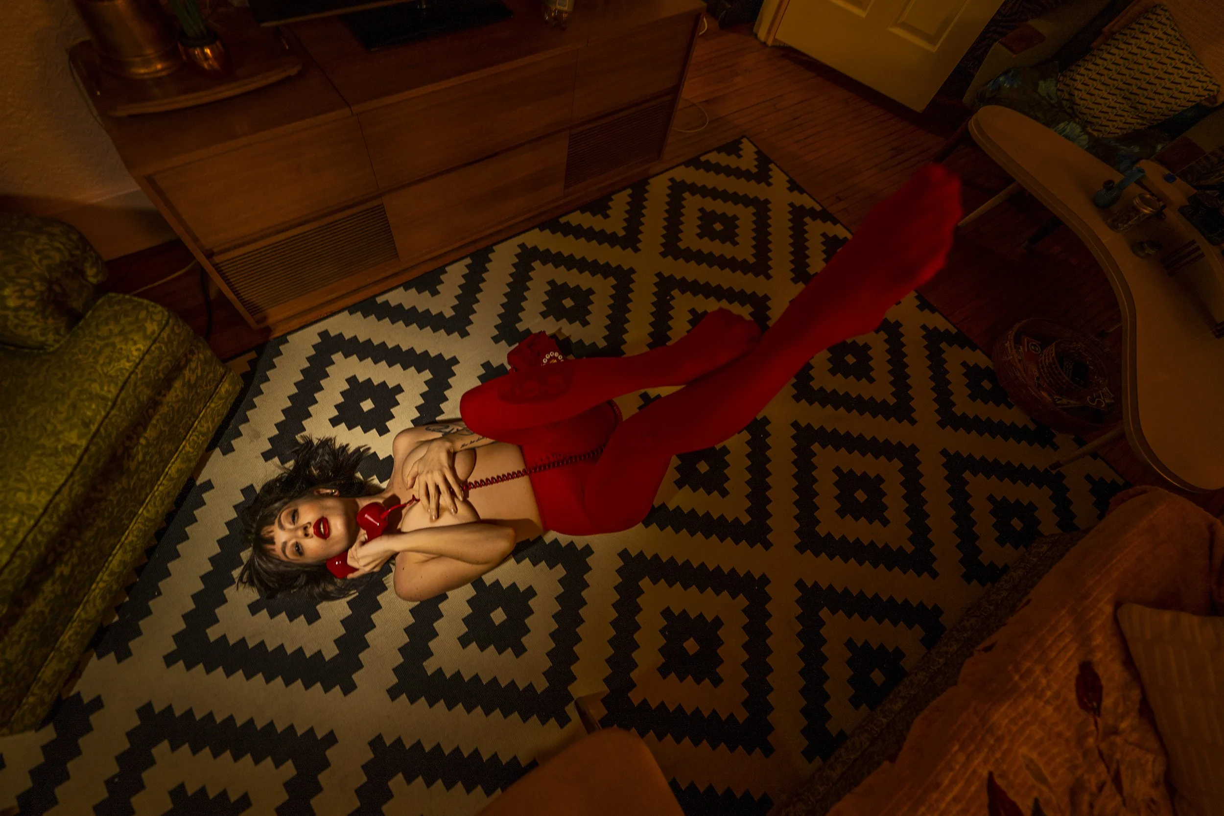 Woman lying on the floor in a red outfit, talking on a red vintage rotary phone, on a patterned black and white rug.