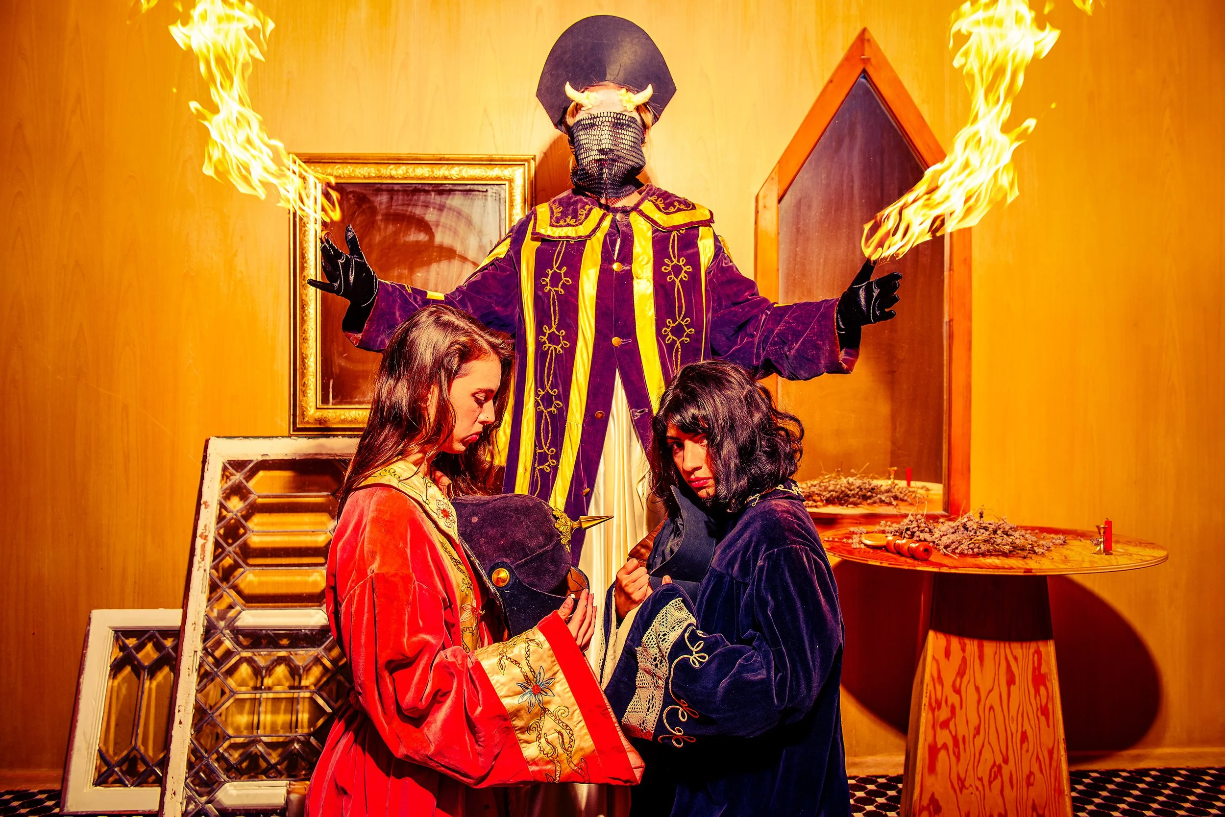 Person wearing a black mask and a costume with horns, standing behind two women with vintage clothing, surrounded by mirrors and flames, in a theatrical or artistic setting.
