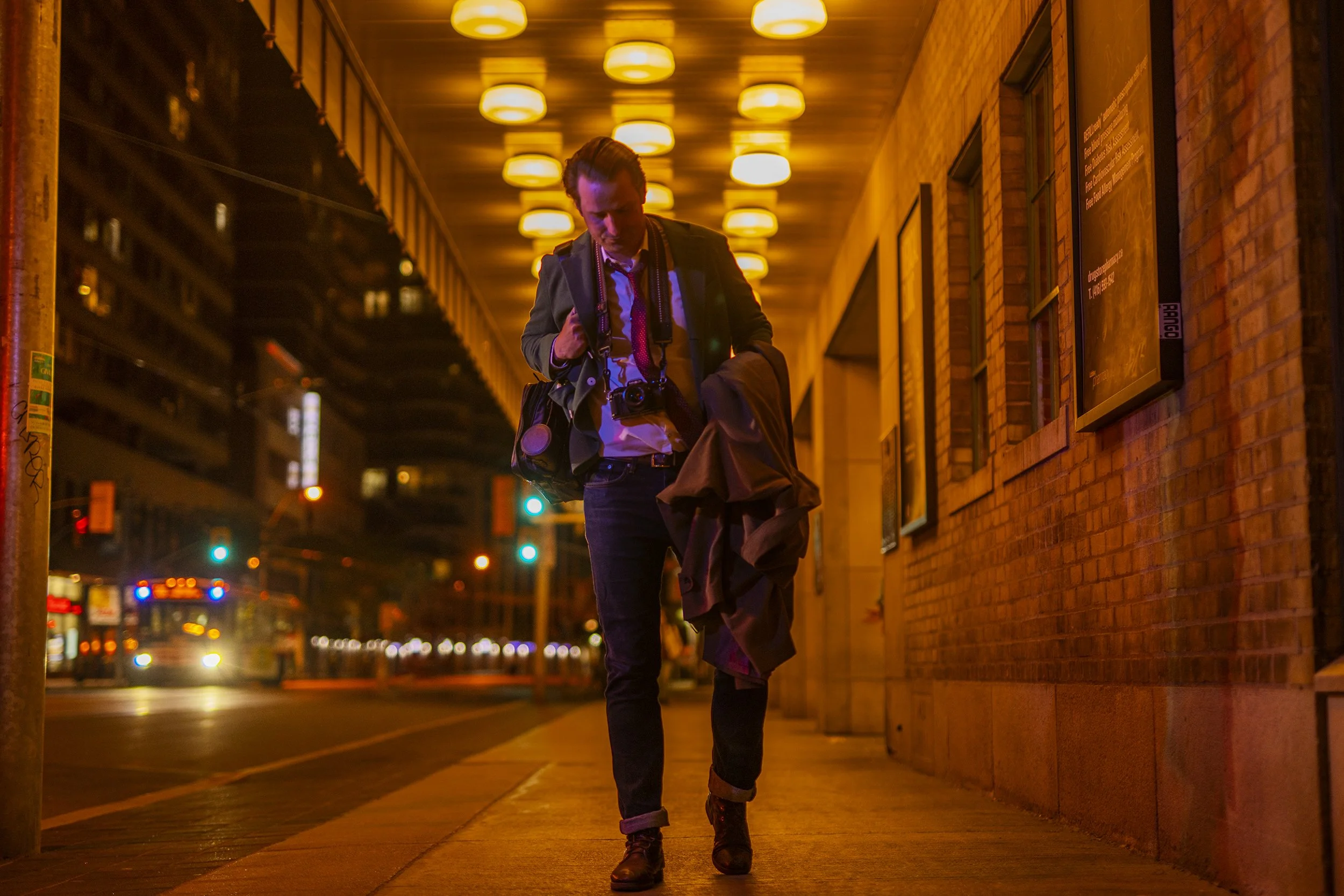 A man with a camera around his neck walking on a city sidewalk at night, with a brick building on one side and city lights in the background.