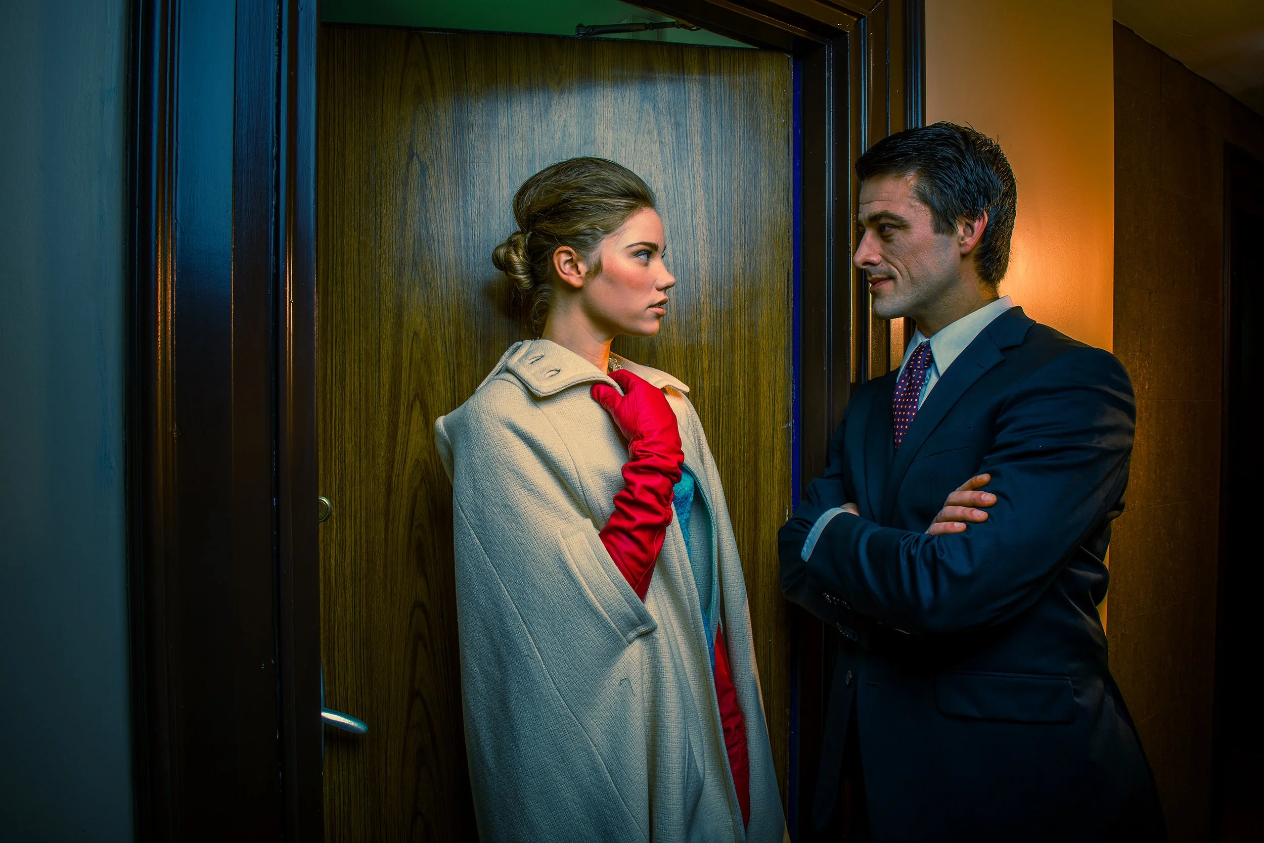 A woman with styled hair and makeup standing in an elevator, looking serious, wearing a beige coat and red gloves. A man in a dark suit with crossed arms and slicked-back hair is facing her and smiling slightly. The elevator has wooden paneling and i