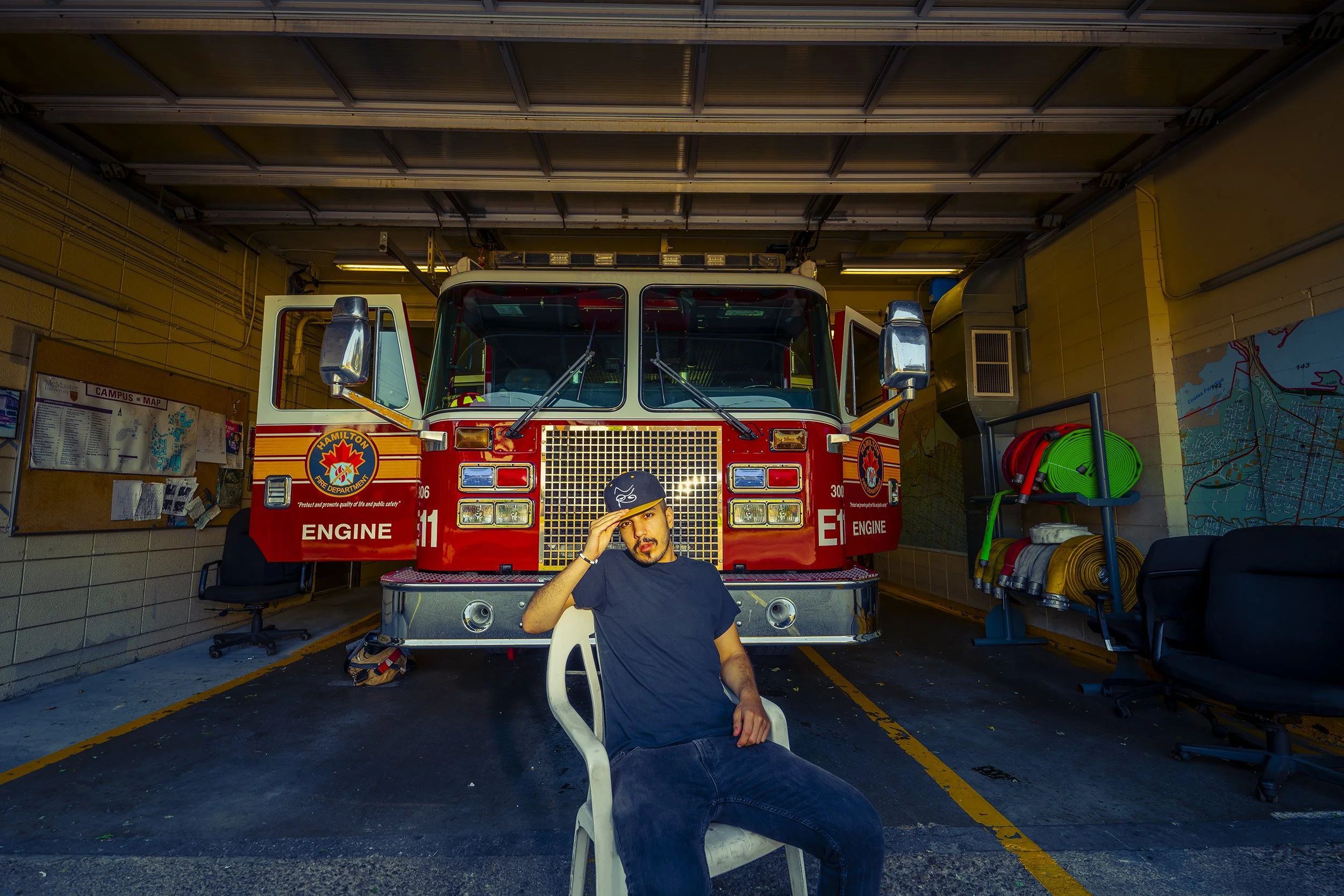 A man sitting in a white plastic chair in front of a parked red fire truck inside a garage. The man is looking at the camera, making a saluting gesture. The garage has a parking lot with yellow lines, bulletin boards, and various equipment including 