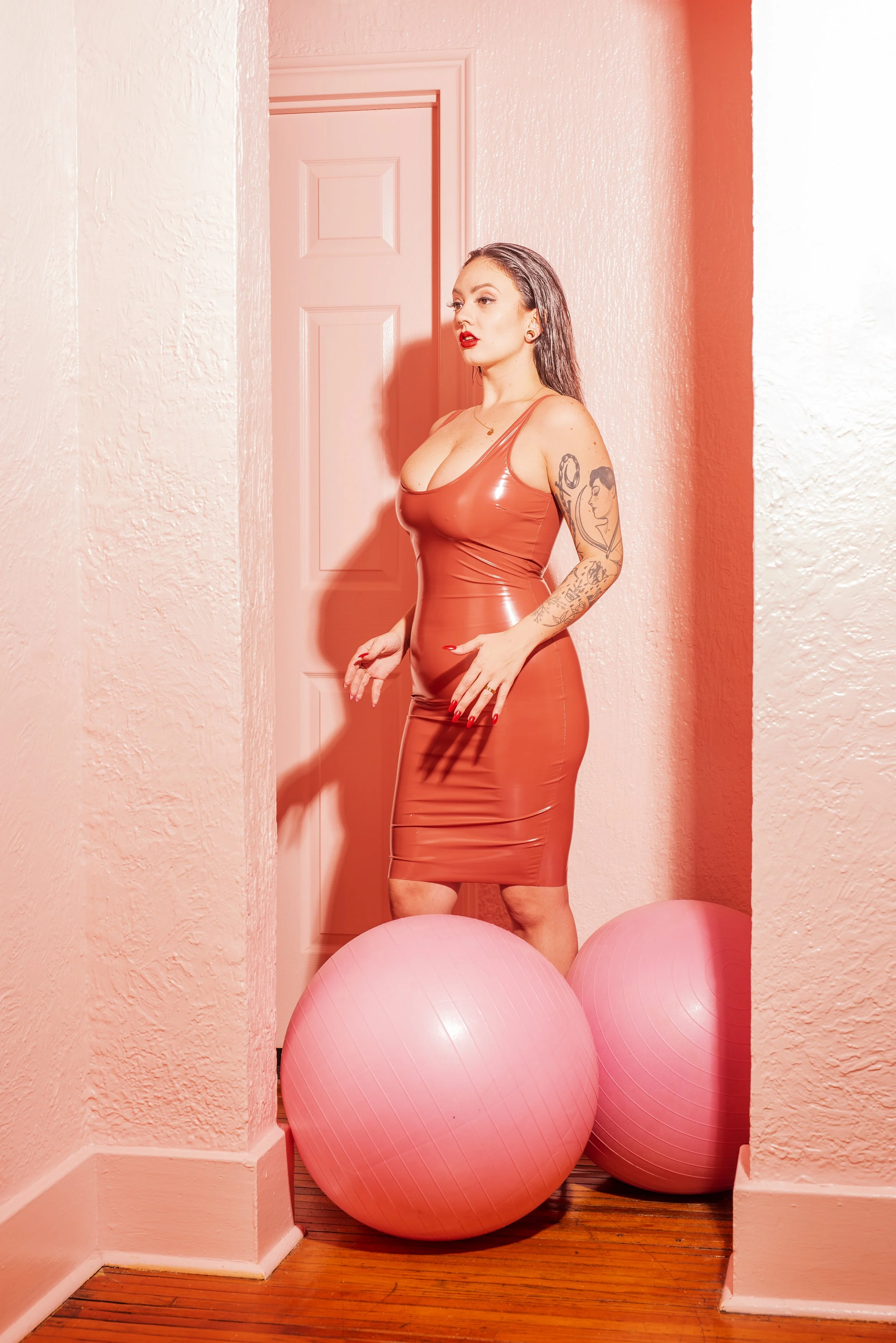 A woman in a red, shiny, form-fitting dress standing between two pink exercise balls in a corner with pink walls, with her hands near her waist.
