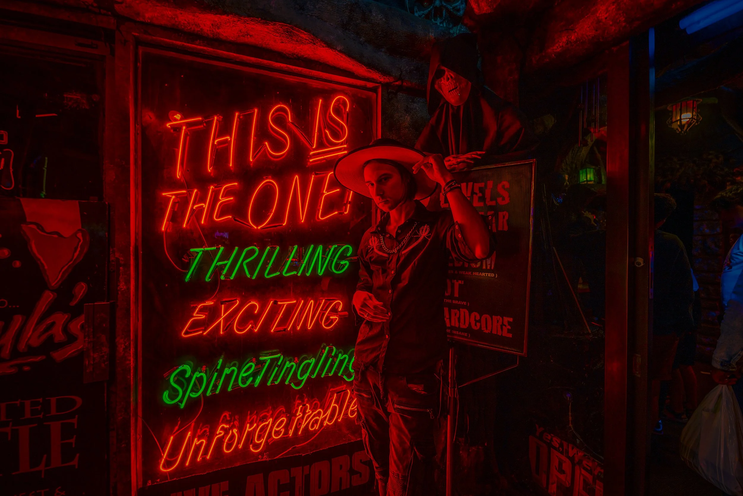 A person wearing a hat and outfit with a skeleton mask standing in front of a brightly lit neon sign that reads 'THIS IS THE ONE!'. The neon sign is red, with some green text below, and the background appears dark and colorful.