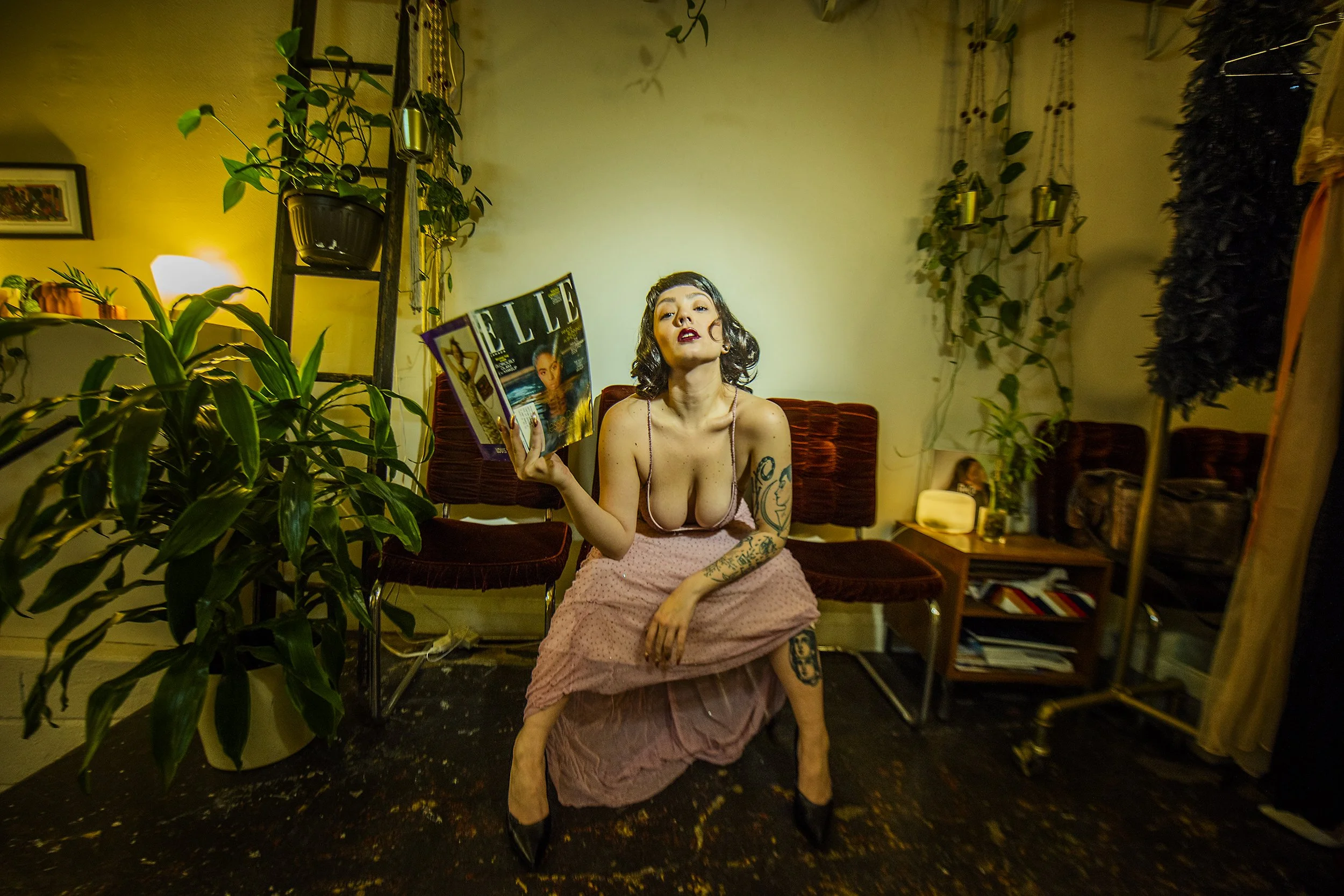 A woman with tattoos sitting on a chair in a room, holding a magazine, with plants and decorative items around her.