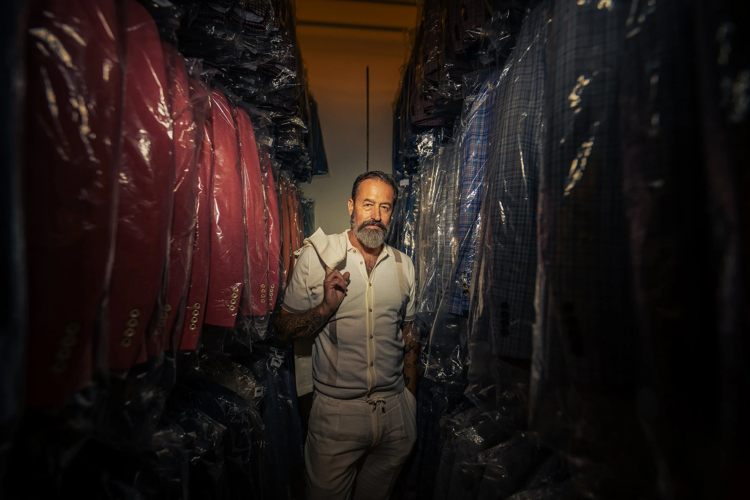 A man with a beard and mustache, wearing a white sleeveless hoodie and beige shorts, carrying a backpack, walking through a narrow aisle in a clothing store with suits and jackets covered in plastic.