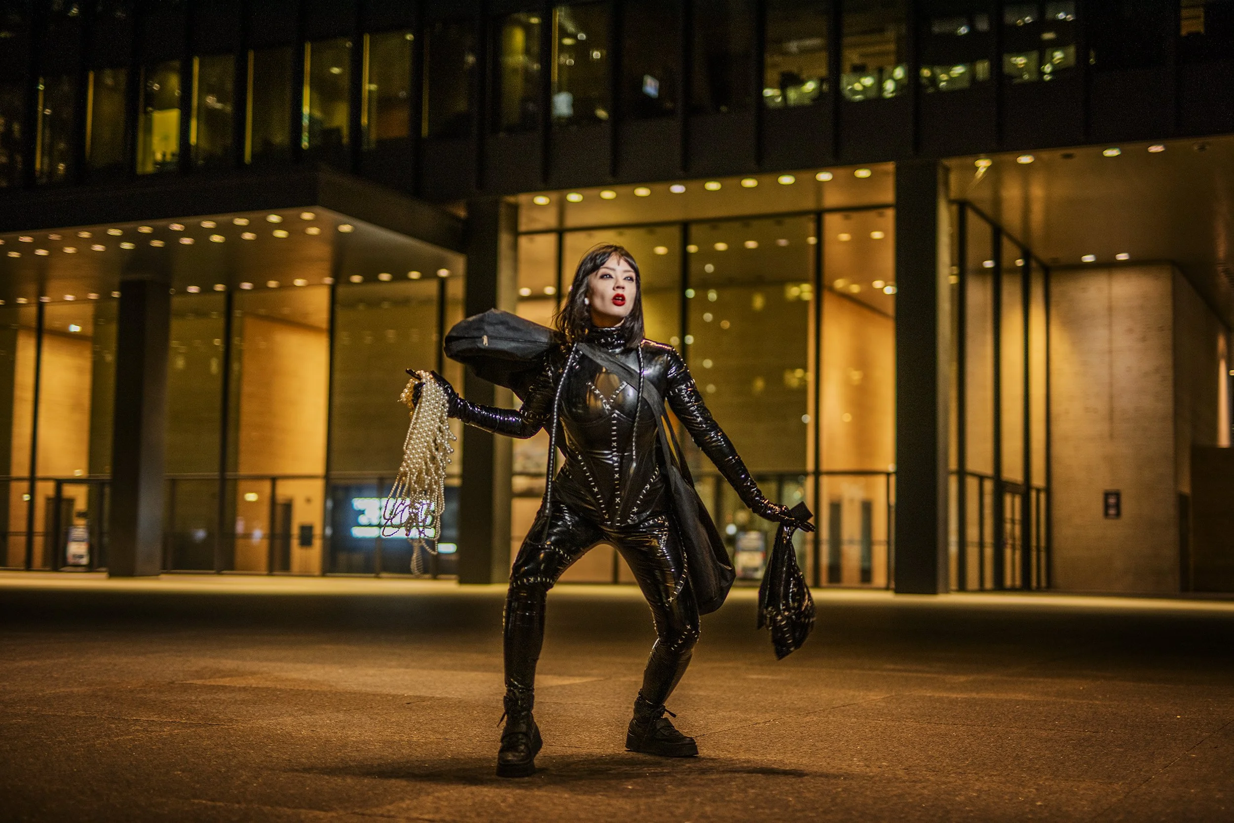 A person with dark hair and red lipstick wearing a black latex outfit standing outside at night, holding a bag with pearl decorations, in front of a modern building with glass windows and warm lighting.
