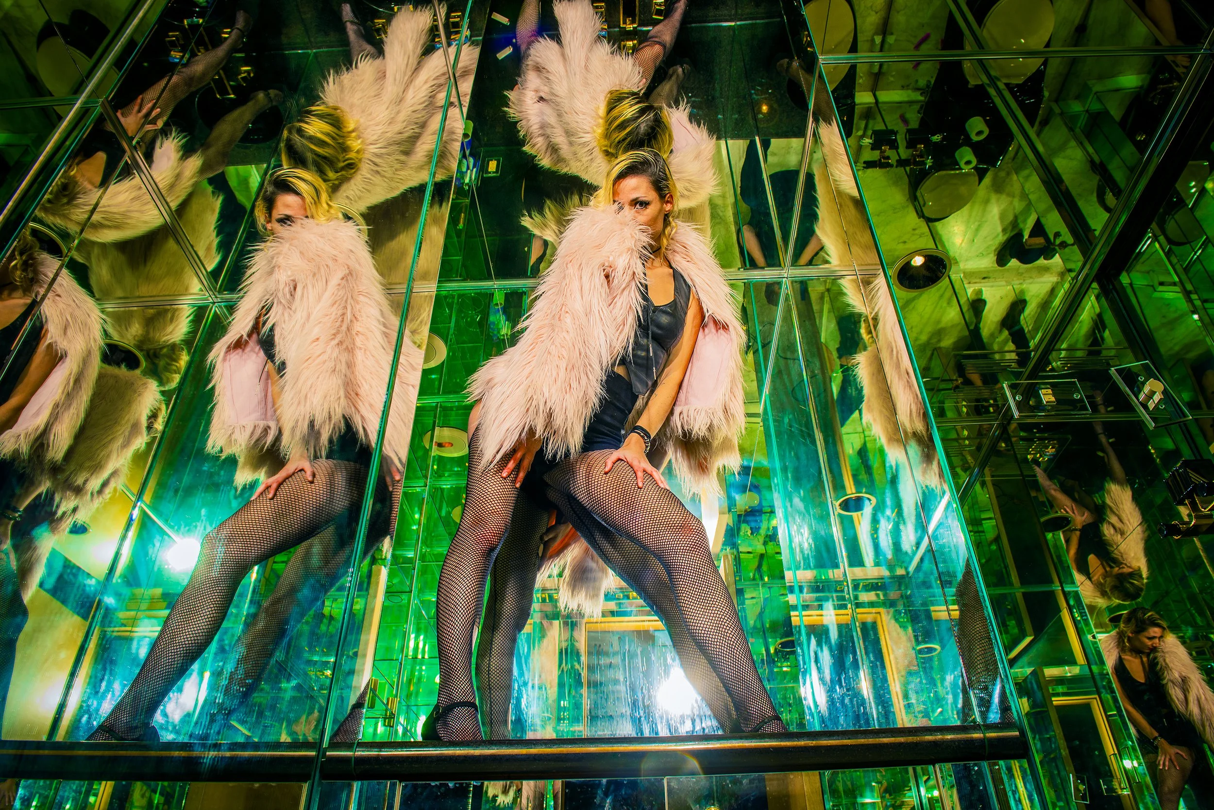 A woman with blonde and black hair posing in a mirrored, green-lit room wearing a pink fur coat, fishnet stockings, and a black leather top, with her hands resting on her knees and looking at the camera.