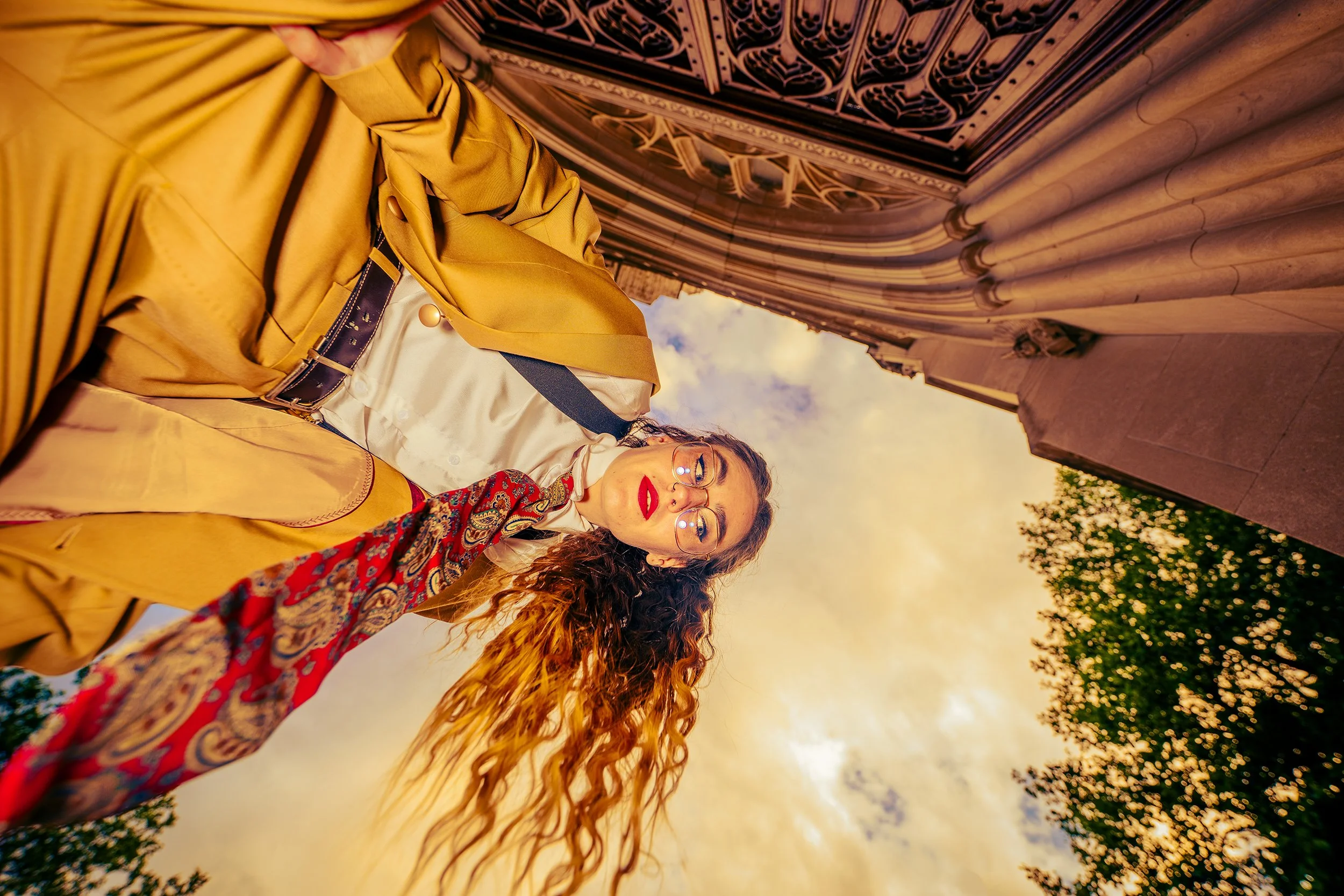 Three women walking outdoors during sunset, with one wearing glasses and a yellow raincoat, another with long red hair wearing a colorful scarf, and the third with curly hair wearing a beige coat. They are near a historic building with columns and in