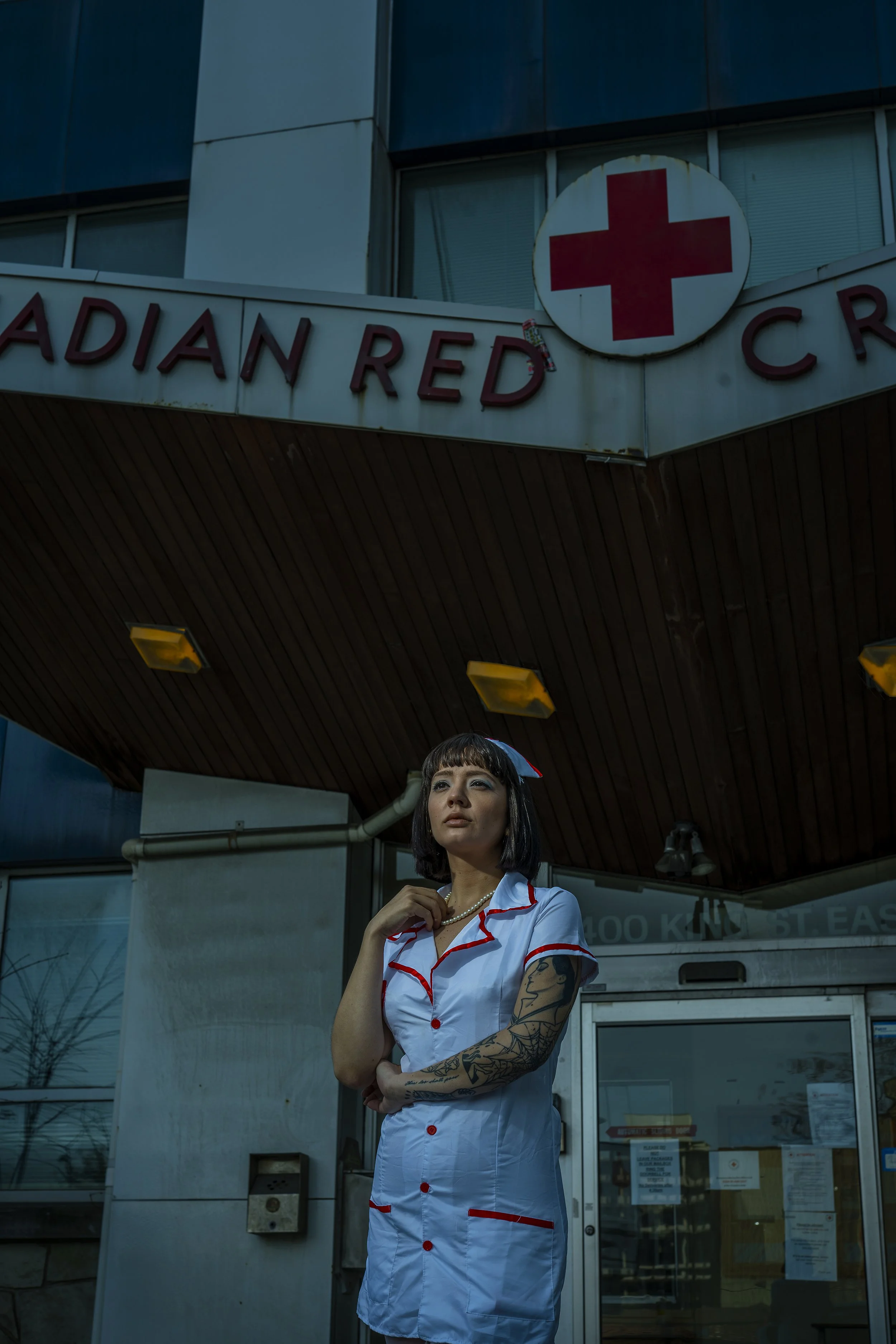 A woman dressed in vintage nurse attire with dark hair and tattoos, standing outside a building with a sign reading "Radiant Red Cross" and a red cross symbol above her.