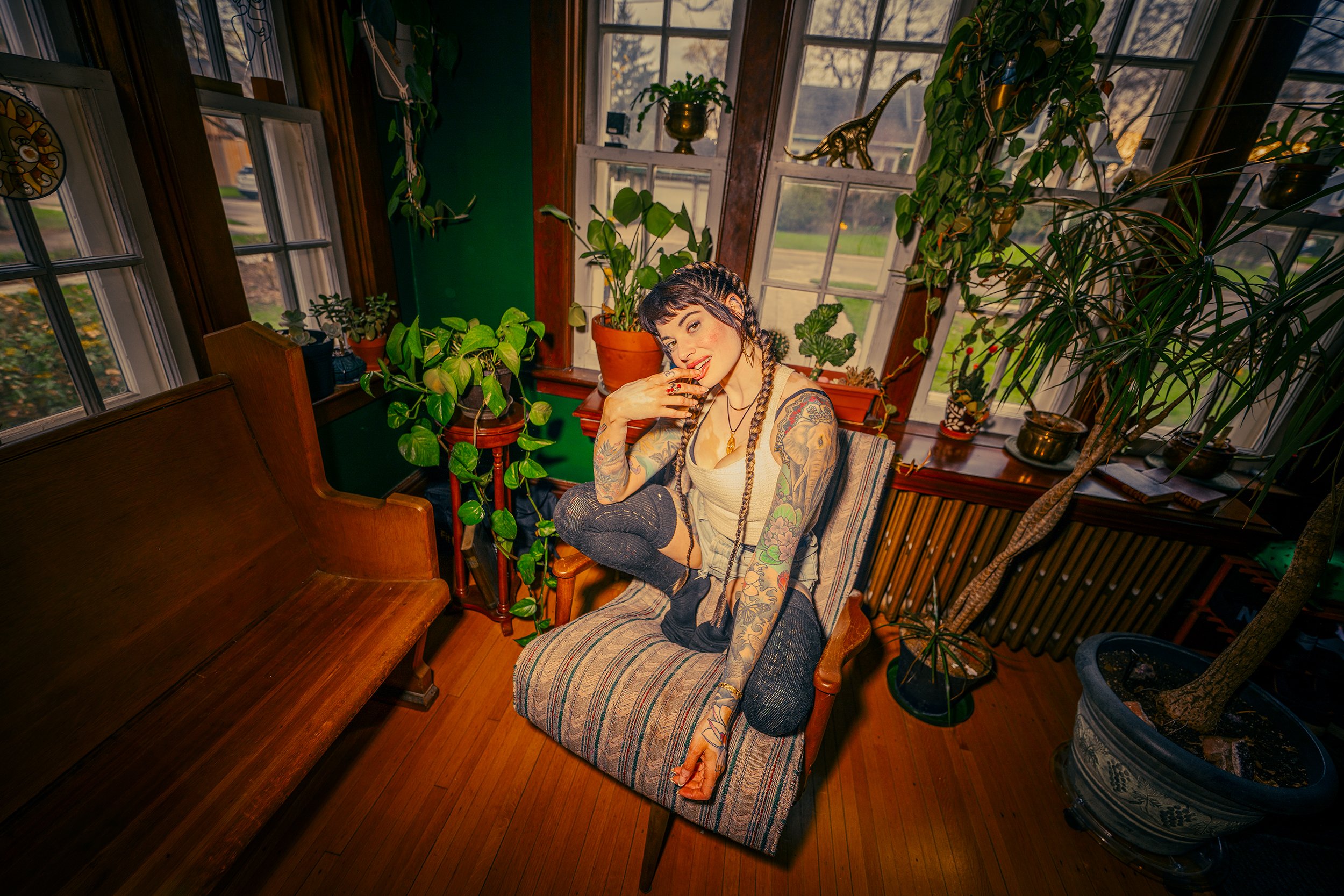 A woman with dark hair styled in braids and tattoos on her arms is sitting comfortably on a striped armchair in a sunlit greenhouse, surrounded by various green plants on window sills and shelves with decorative objects.
