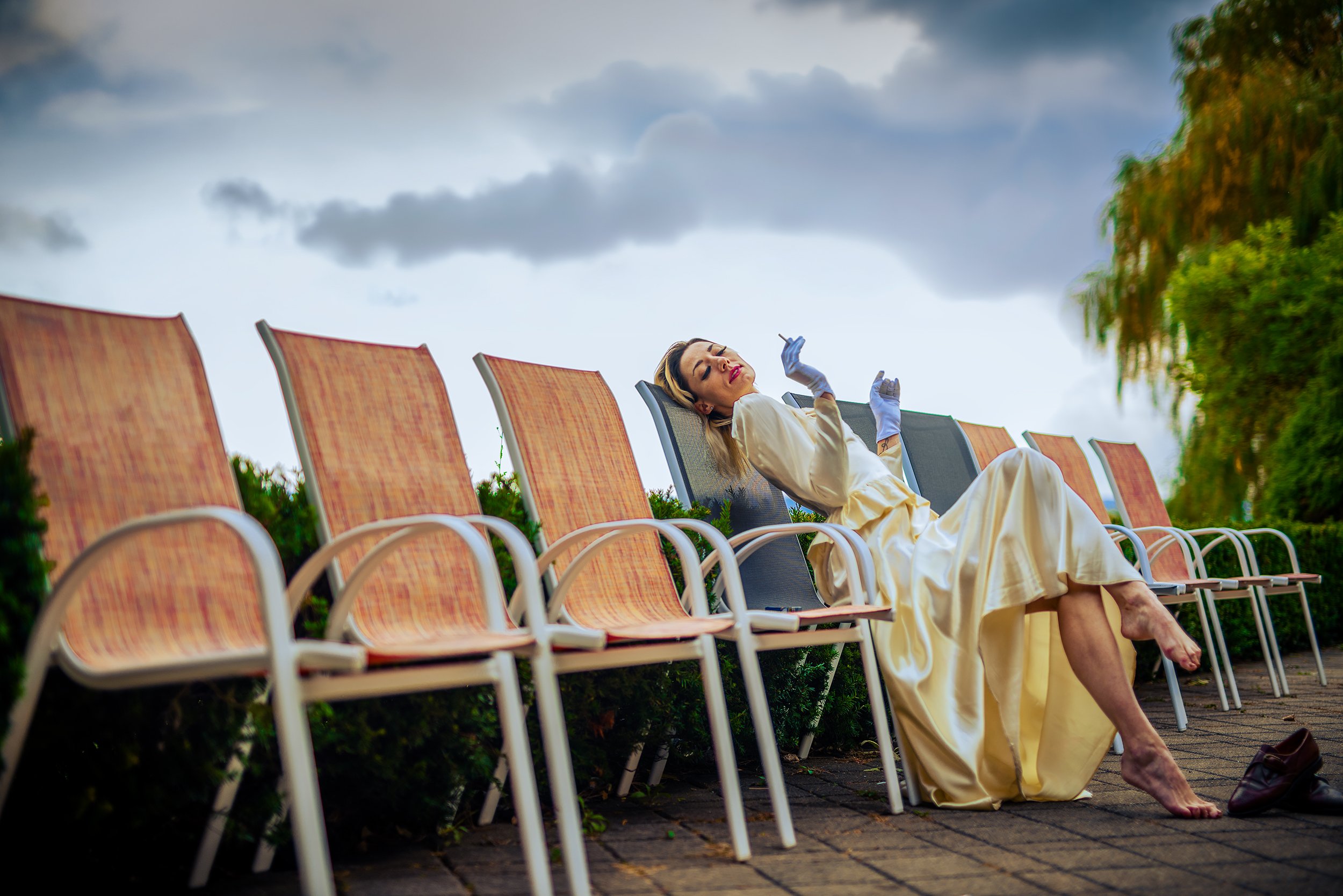 A woman in a yellow gown sitting on a row of orange chairs outdoors, leaning back with eyes closed, wearing blue gloves and holding a cigarette, with overcast sky and trees in the background.