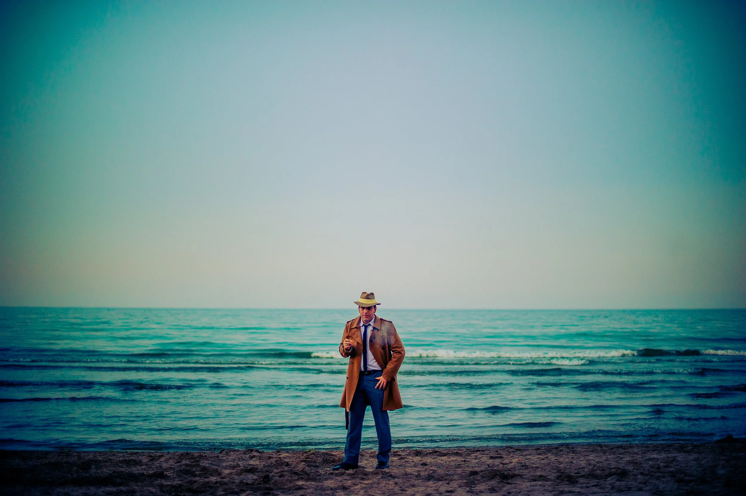 A man in a brown trench coat, hat, and tie stands on a beach with the ocean behind him. He is smoking a cigarette and looking at the camera.