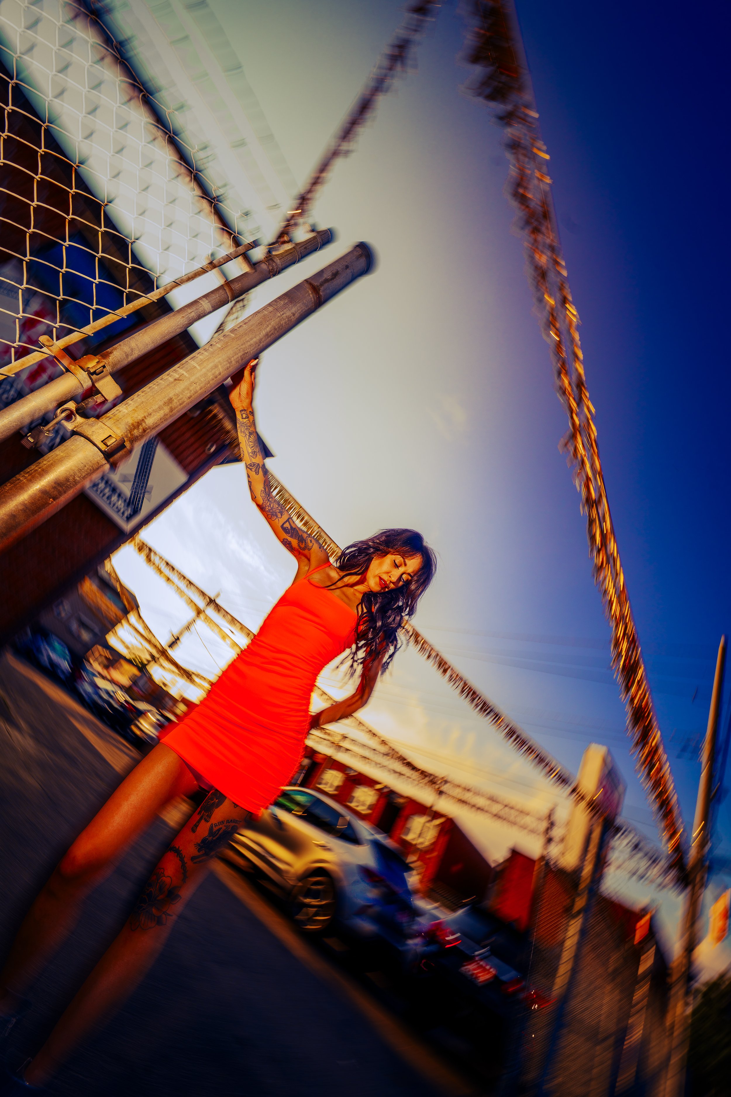 A woman in a red dress standing outdoors near a fence with arms raised, at sunset with a Ferris wheel in the background
