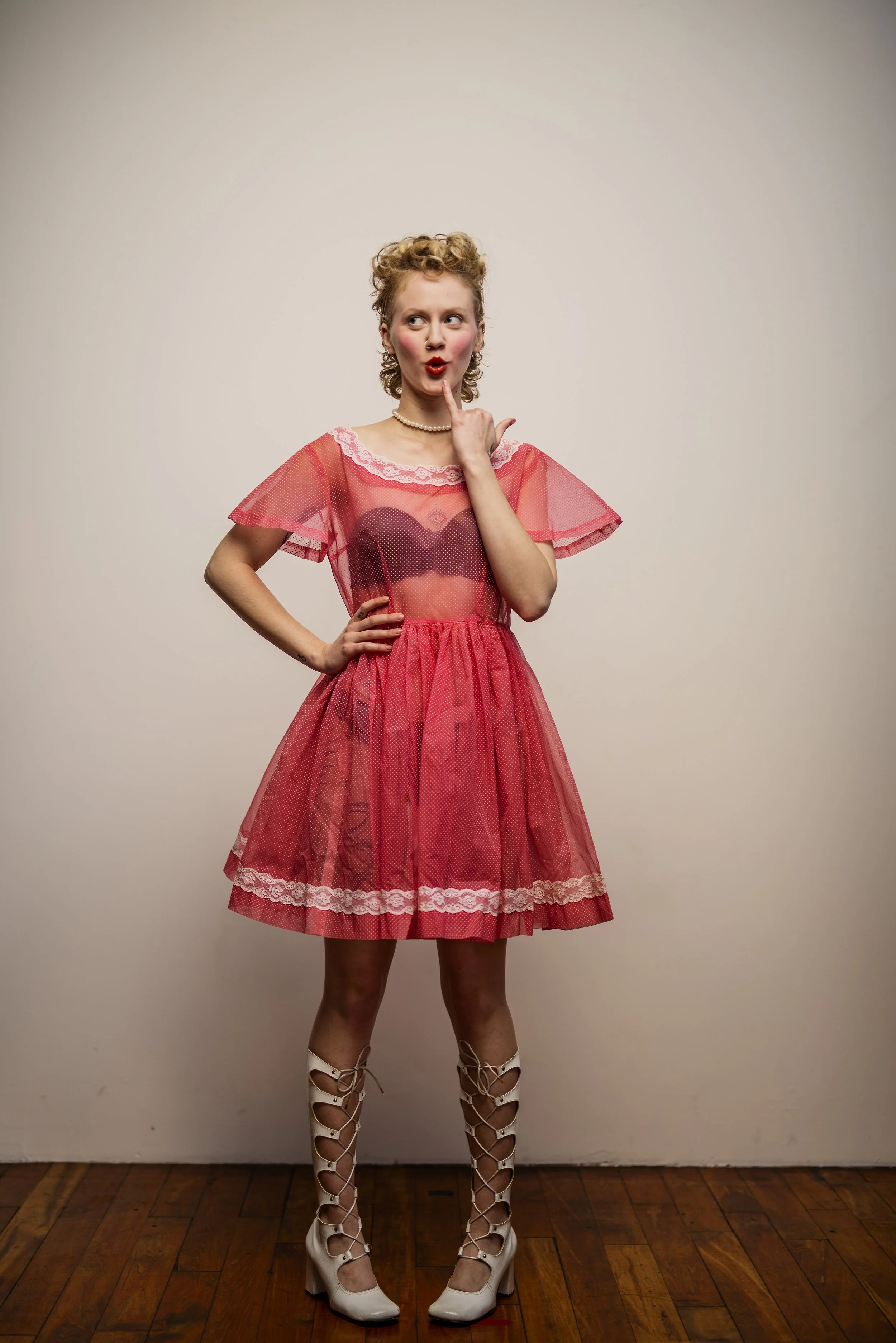 Woman in a pink dress with lace details and mesh overlay, wearing white lace-up high-heeled shoes, standing on wooden floor against plain wall.