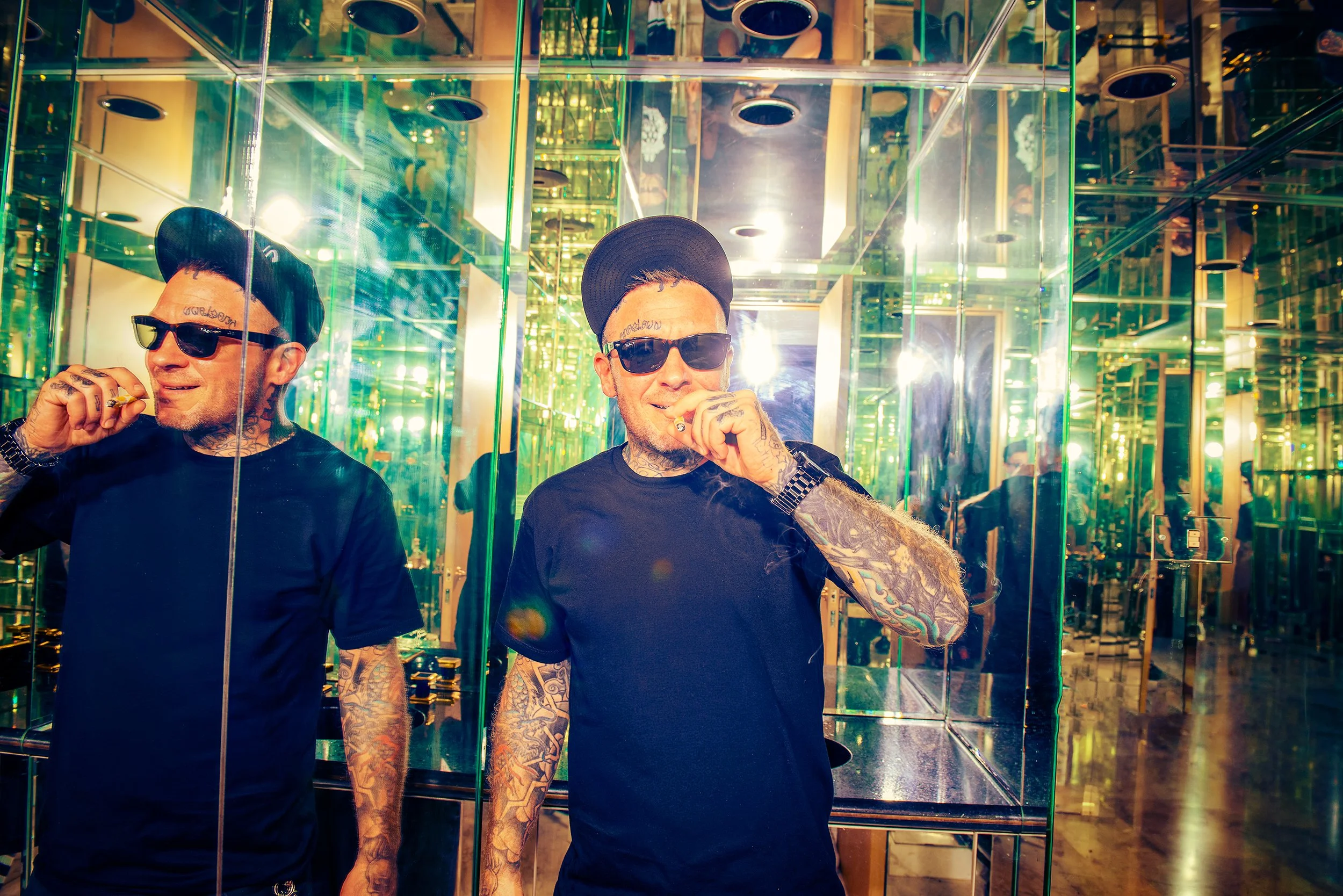 Two tattoos men in black shirts and black hats, one with sunglasses, are inside an elevator with mirrored walls, reflecting lights and the interior design.